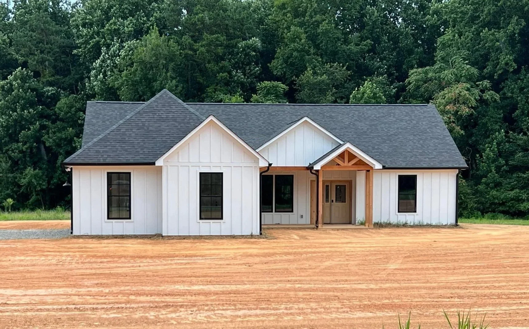 New house under construction with white exterior walls, black window frames, and a dark gray roof, set on a dirt lot with green trees in the background.