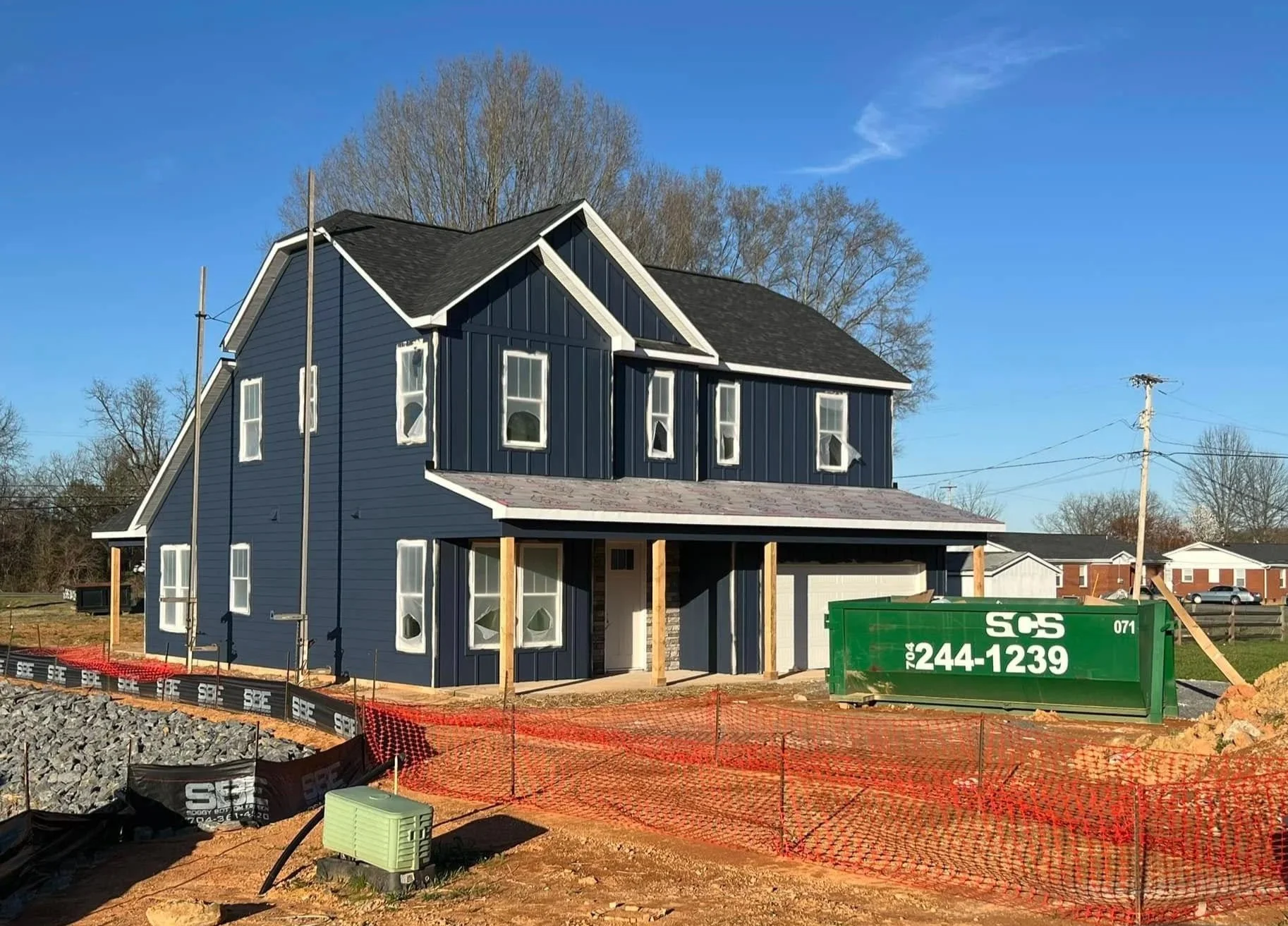Two-story house under construction with blue siding, white trim, and a black roof, surrounded by construction materials and equipment, with a green dumpster nearby, under a clear blue sky.