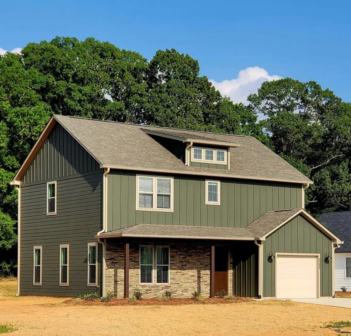 New three-story house with green siding, stone accents, and a one-car garage, surrounded by trees under a blue sky.