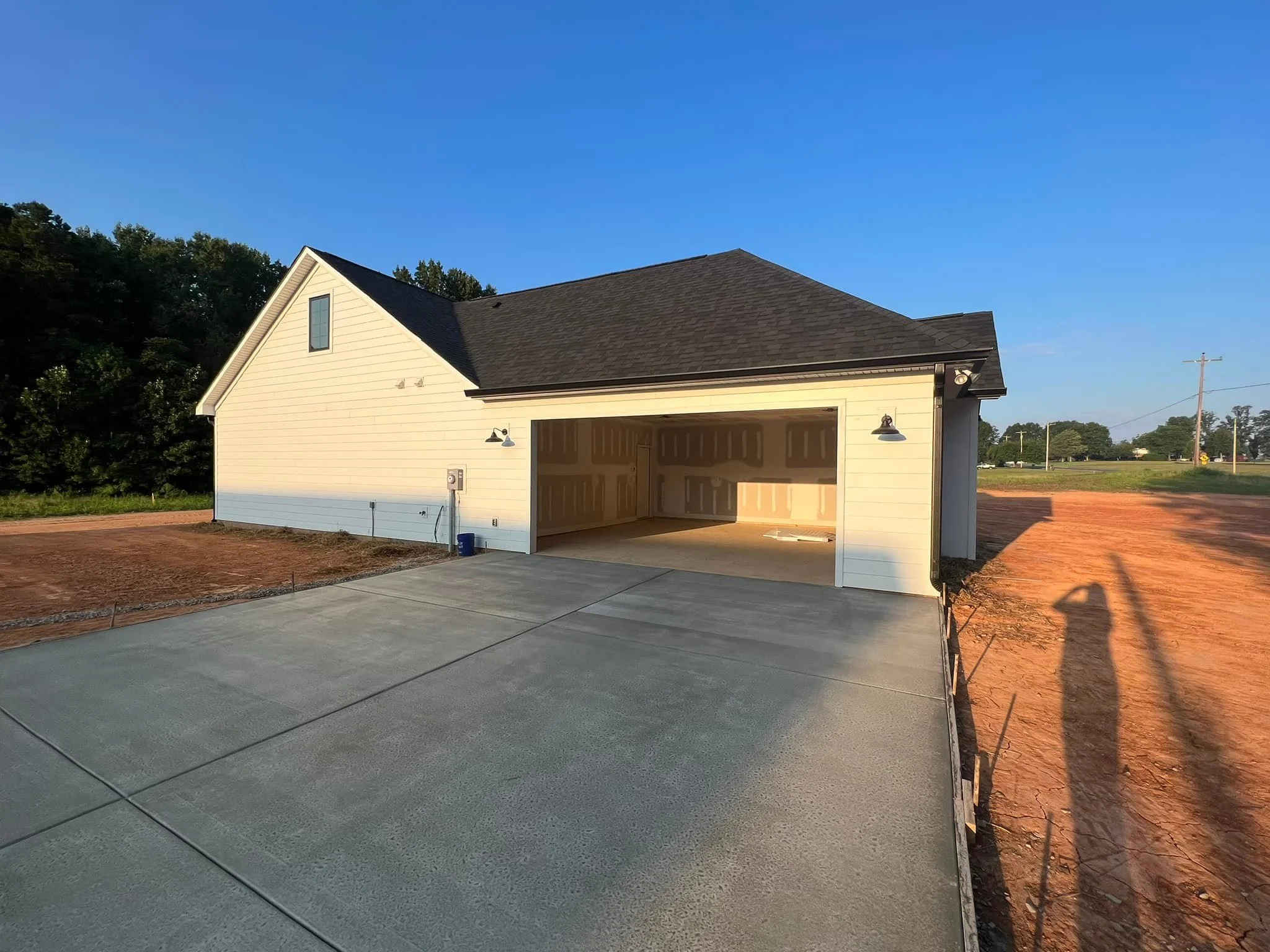 Newly built detached garage with a concrete driveway, white siding, black roof, and exterior lighting in a suburban area, with a blue sky and green trees in the background.