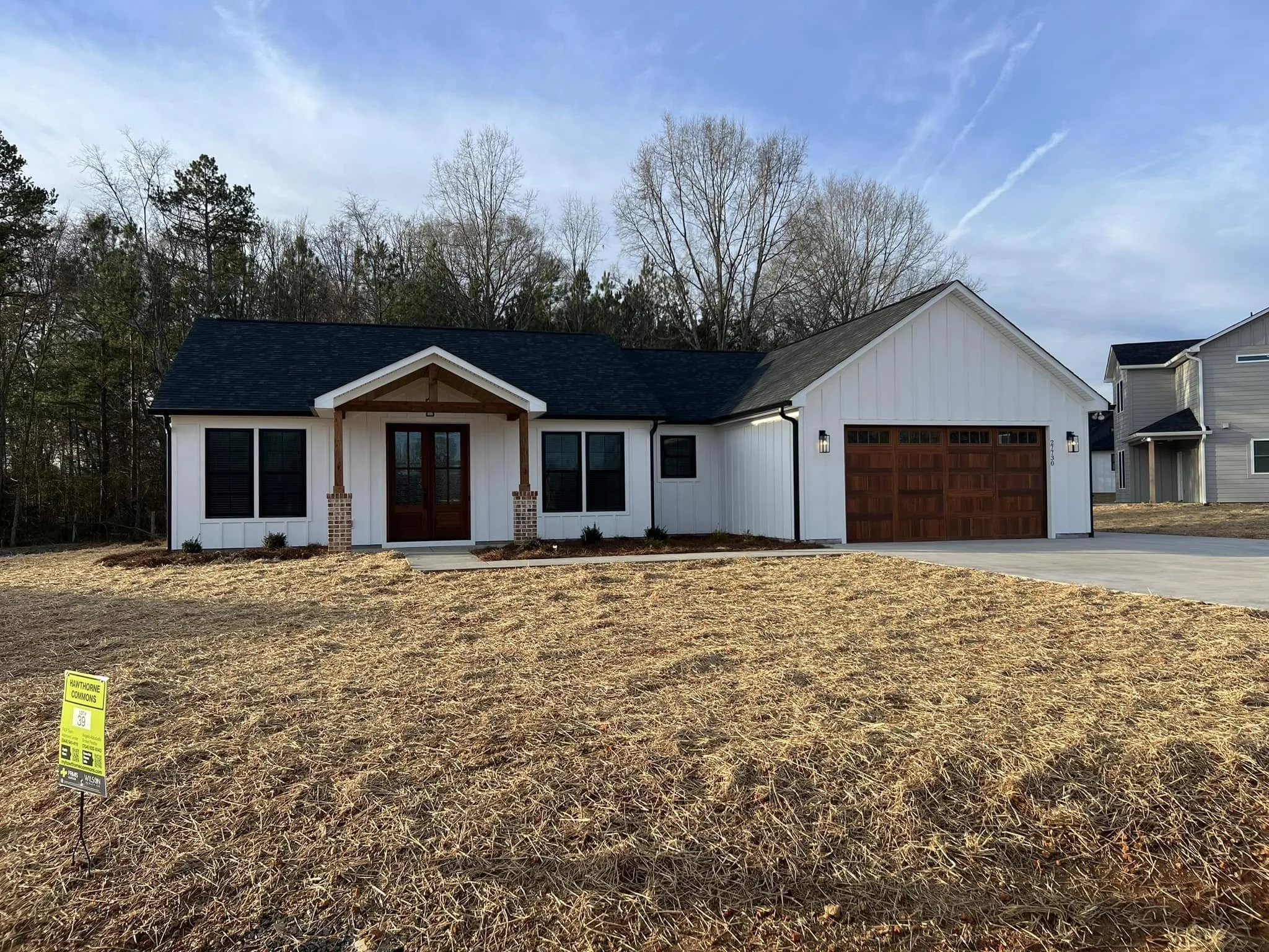 Front view of a modern white house with a dark roof and wooden garage door, new landscaping, and a small front porch with two brick columns. A yellow construction sign is in the yard.