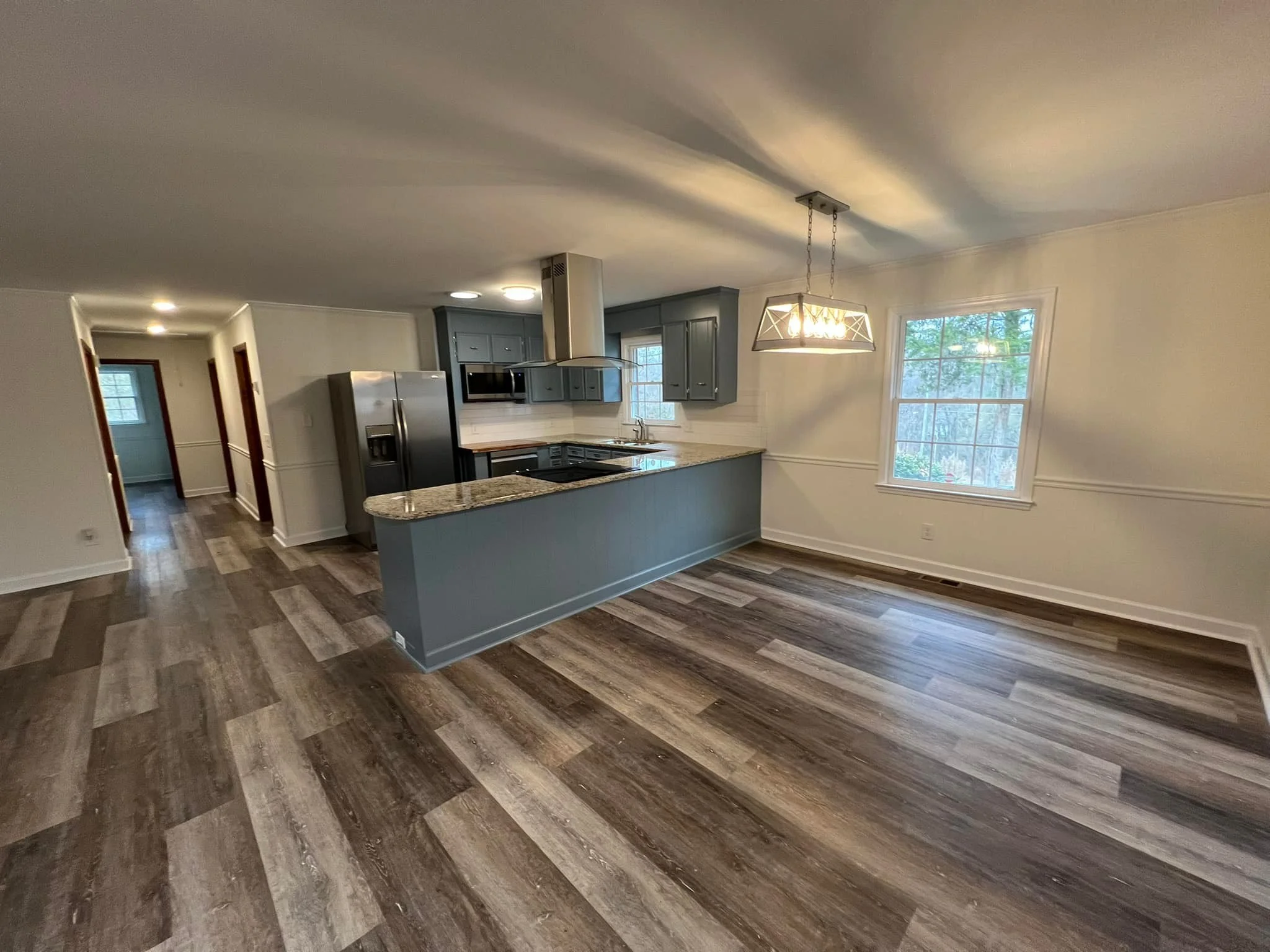 Empty modern kitchen and dining area with gray cabinets, stainless steel appliances, granite countertops, wood-look flooring, and a large window, with a hanging light fixture.