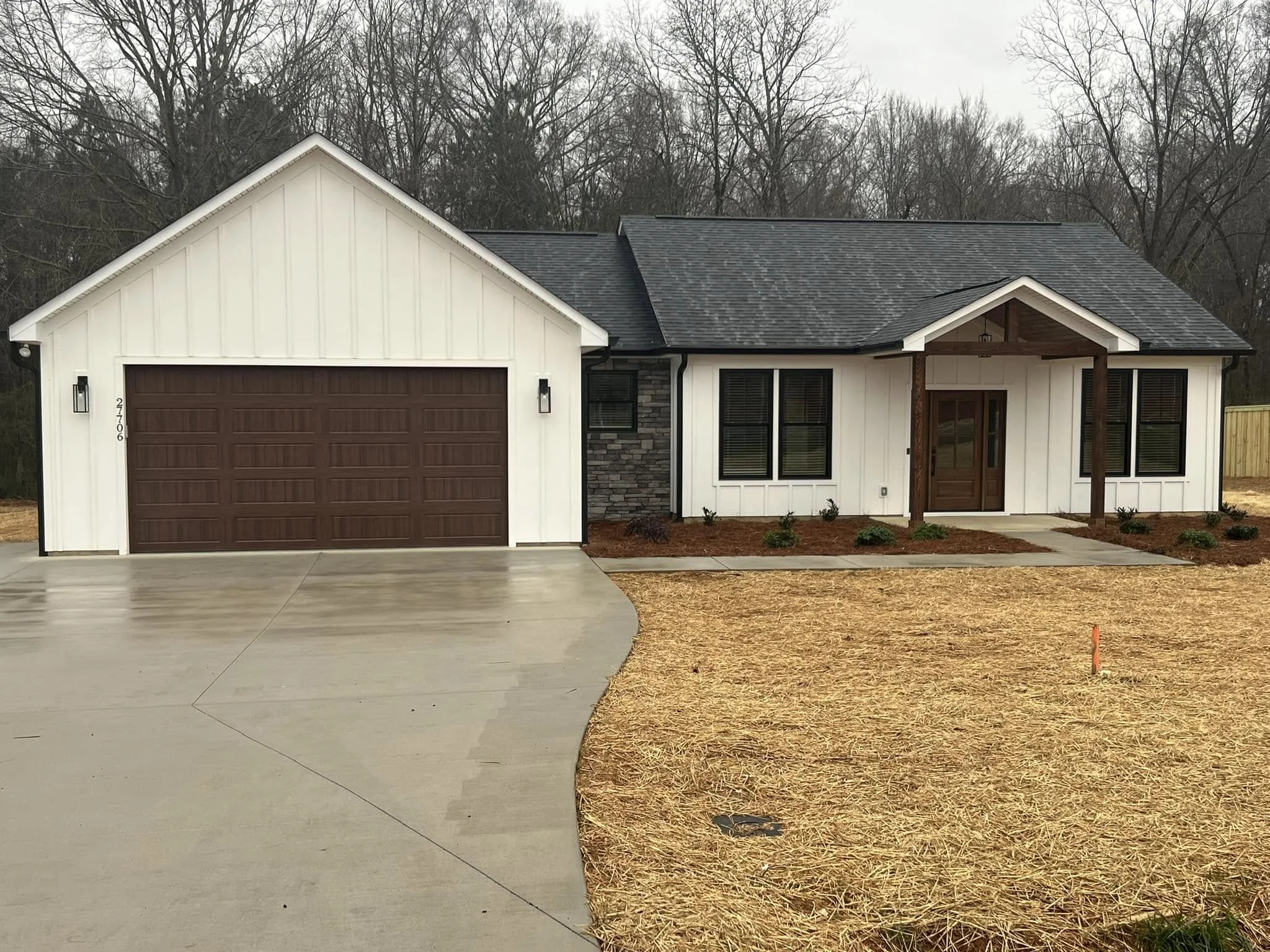 Single-story modern house with white exterior, brown garage door, front porch with wooden beams, and landscaped yard with small bushes and mulch.