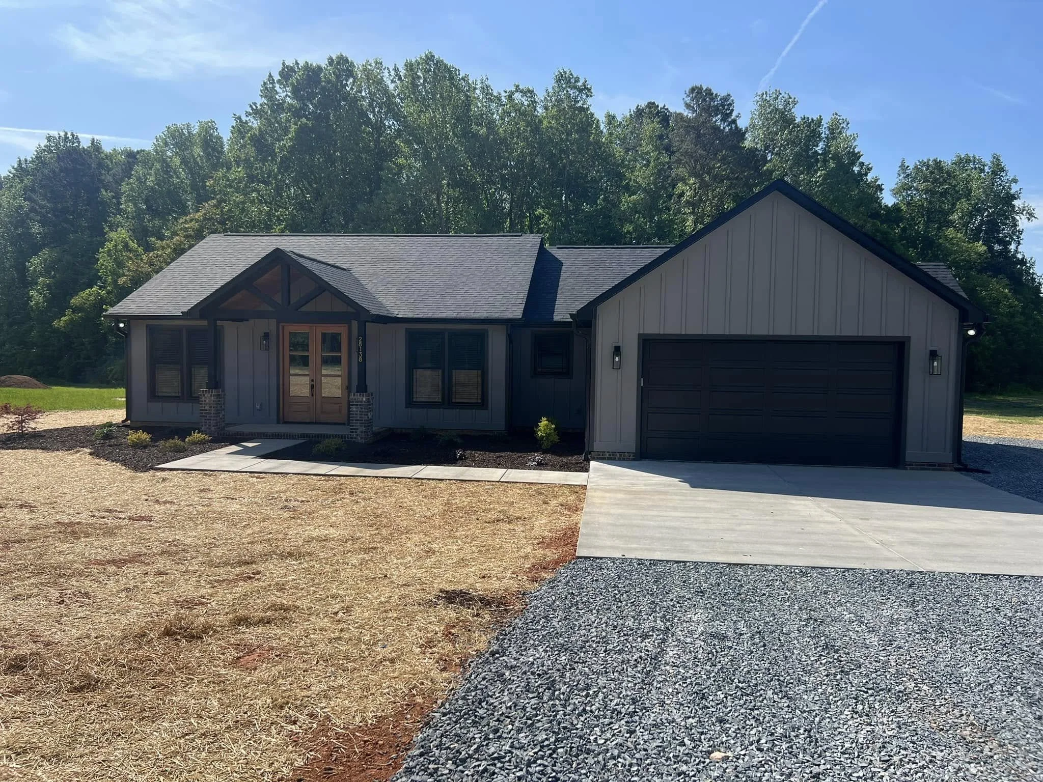 Front view of a newly built house with gray exterior, black garage door, front porch with wooden door, surrounded by a new gravel driveway and a small landscaped area, with a backdrop of green trees under a blue sky.