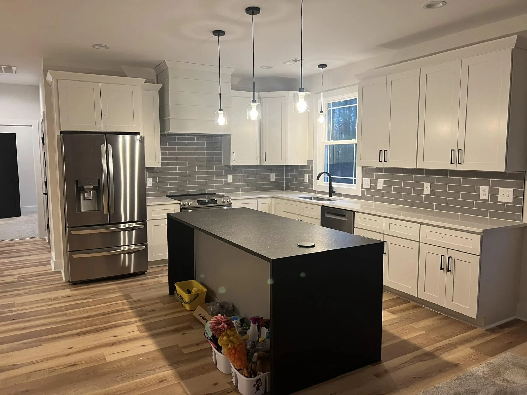 Modern kitchen with white cabinets, stainless steel refrigerator, gray backsplash, black island, and hardwood floors.