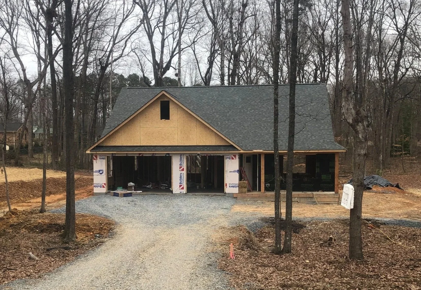 Under construction house with a gravel driveway, surrounded by leafless trees, and building materials visible around the structure.