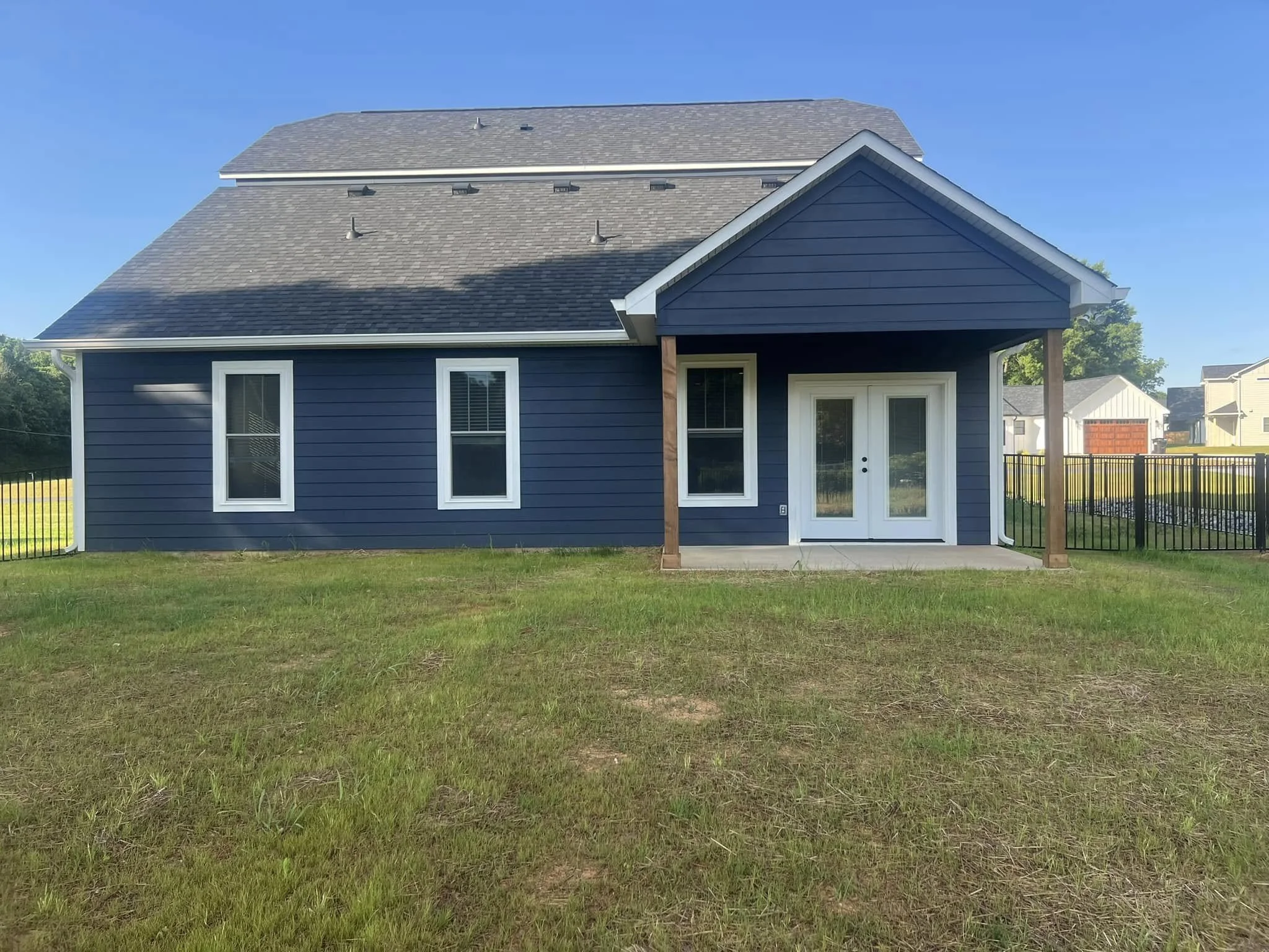 A blue house with white trim, two windows, and a small porch with a white door, surrounded by a grassy yard and black metal fence, with other houses in the background