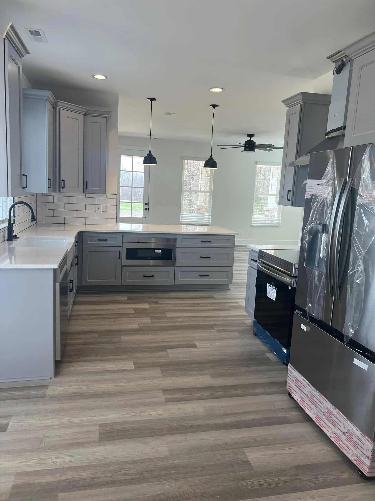 Modern kitchen with gray cabinets, stainless steel appliances, a white countertop, a brick-style backsplash, and light wood flooring, illuminated by pendant lights and natural light from windows.