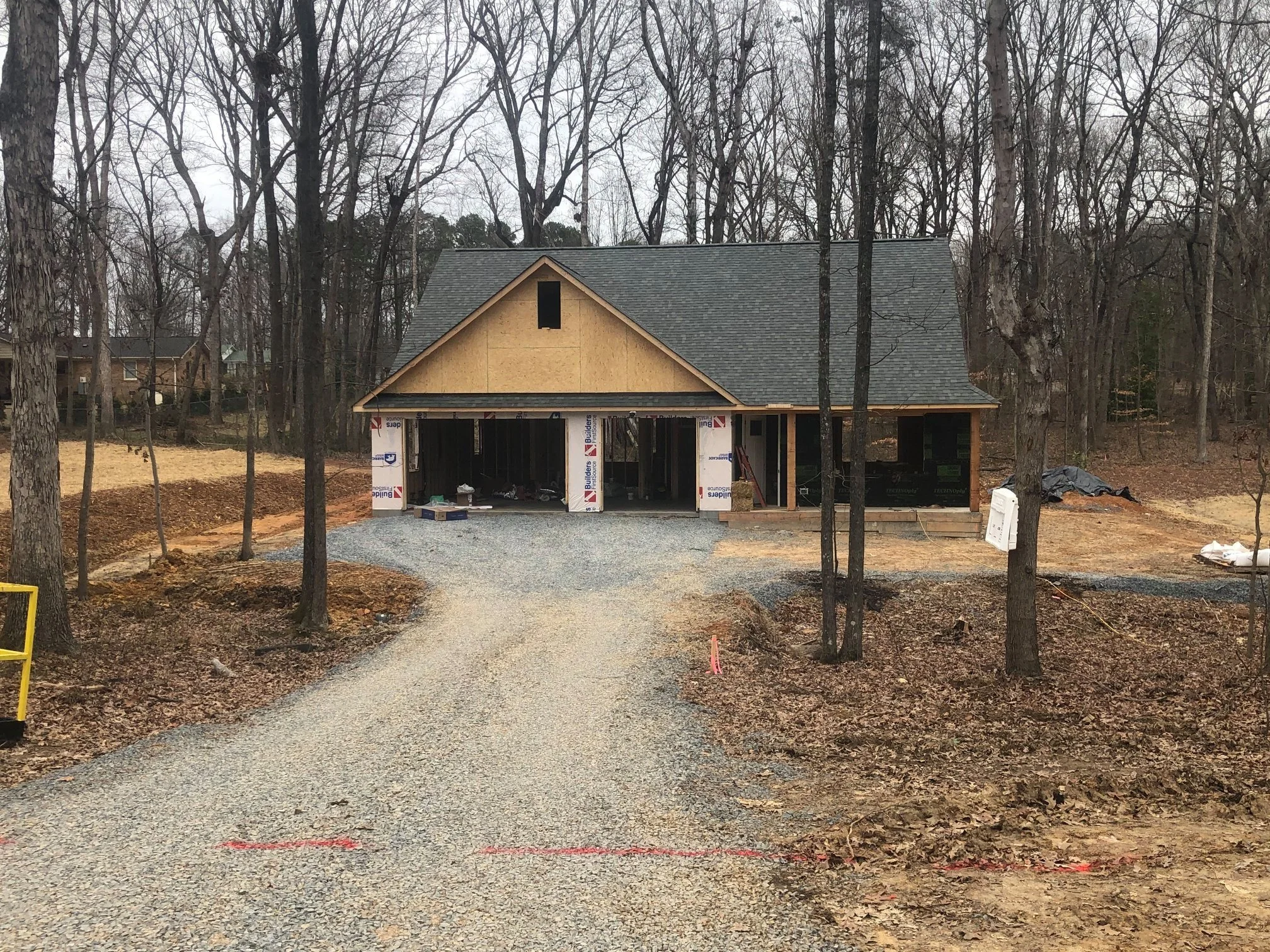 Construction site of a house with an incomplete garage, surrounded by trees and dirt, with building materials and equipment scattered around.