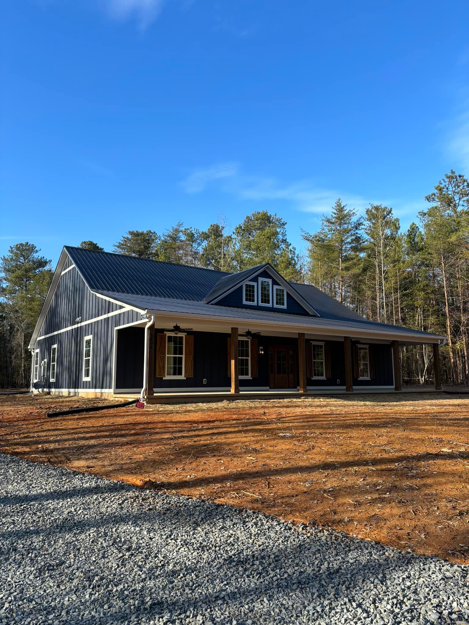Newly built house with dark blue exterior, white trims, and wooden shutters, situated in a wooden area with a gravel driveway and a clear blue sky.
