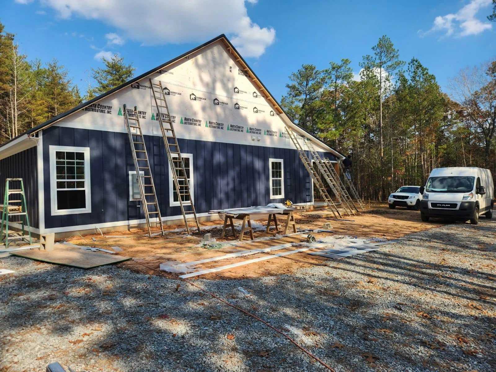 A house under construction with blue siding, scaffolding, and construction tools, surrounded by trees and parked vehicles.