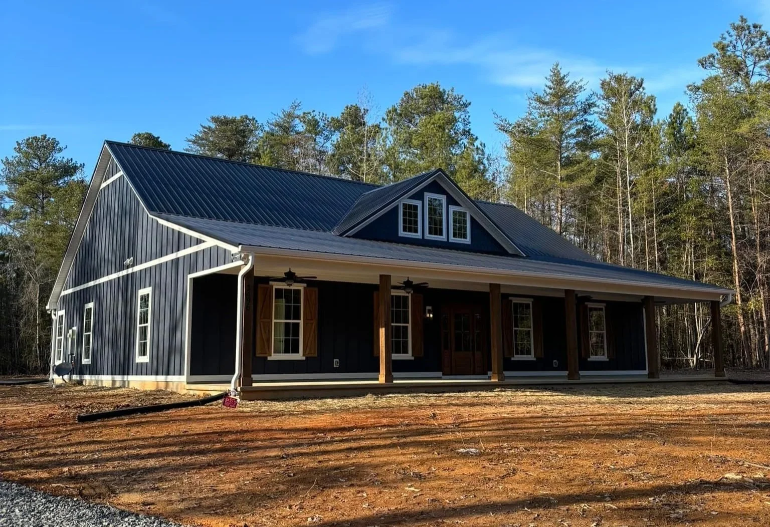 A modern house with black siding, a metal roof, and a covered porch with wooden support beams, set against a background of trees and a blue sky.