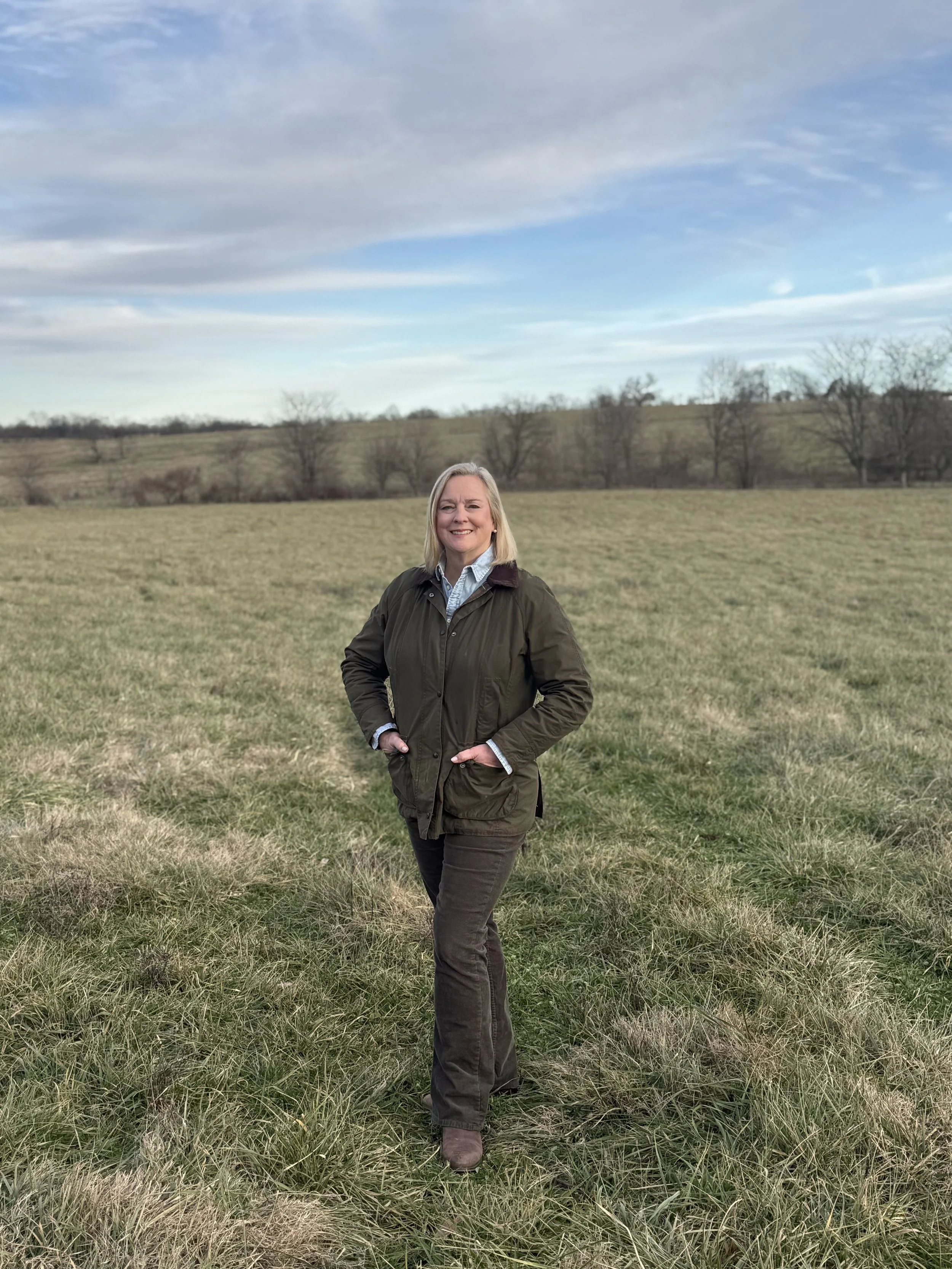 A woman with blonde hair wearing a green jacket and brown pants, standing in a grassy field with a few trees and a cloudy sky in the background.