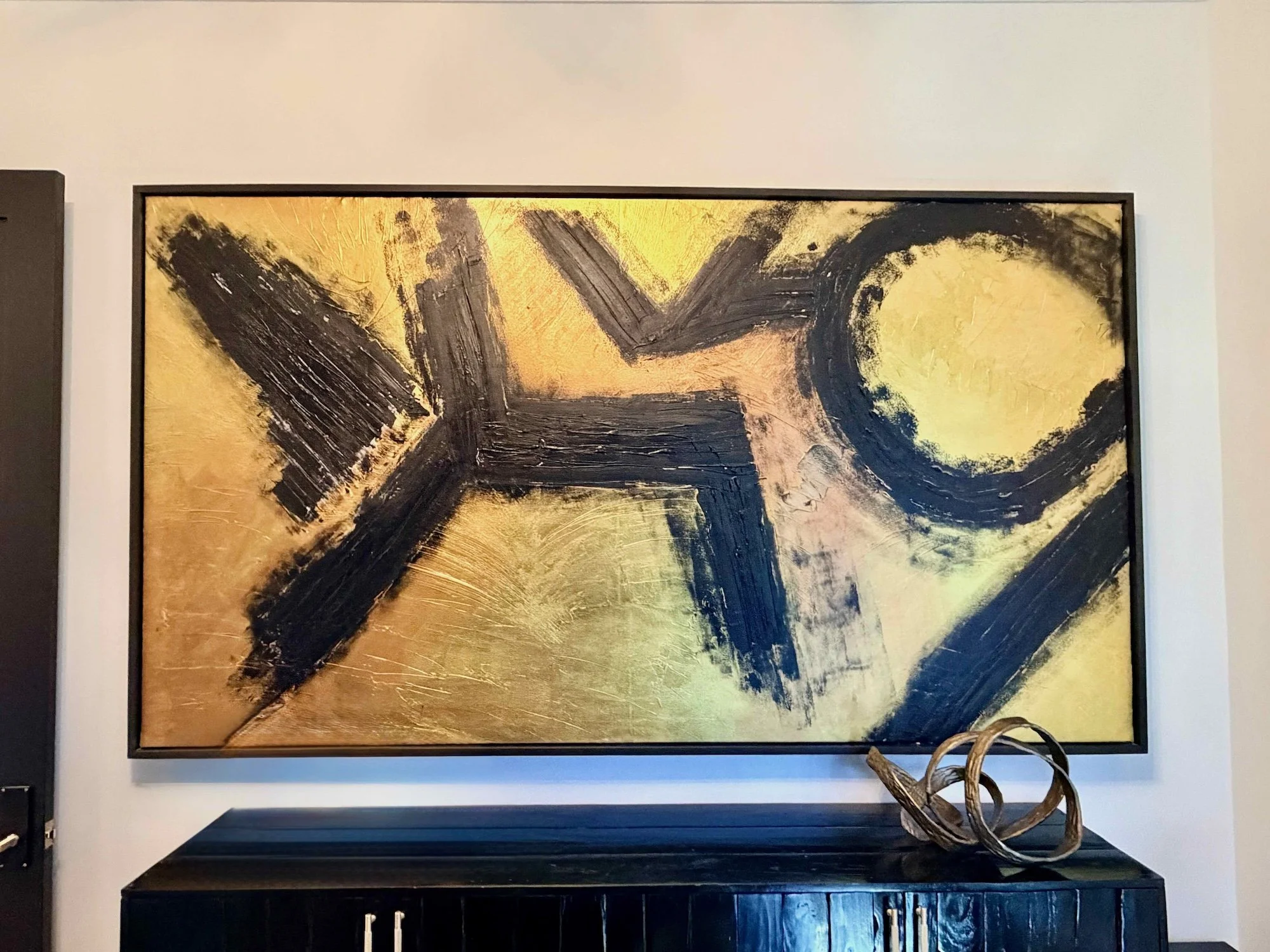 An abstract painting with bold black brushstrokes on a gold and beige background, featuring a circular shape and lines, framed on a white wall above a black console table with abstract sculptures.