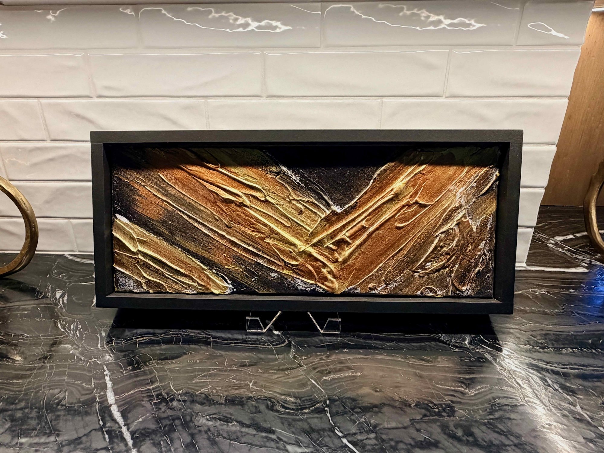 Decorative rectangular fire pit with a textured gold, bronze, and black flame pattern, placed on a black marble surface in front of a white tiled wall.