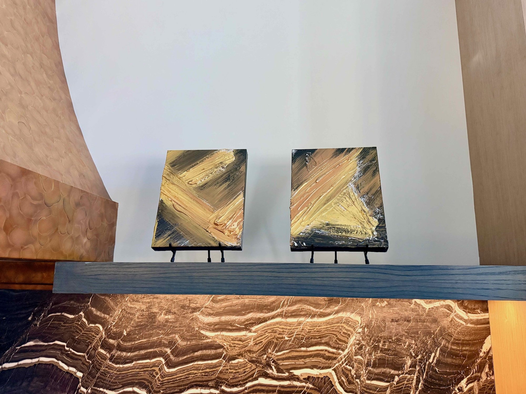 Two abstract paintings on small easels displayed on a dark stone or marble surface with a white wall background.