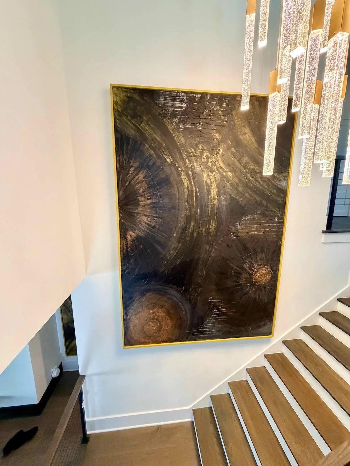 A large abstract painting with dark swirling patterns and circular textures, framed in gold, hanging on a white wall next to staircase with wooden steps and white risers.