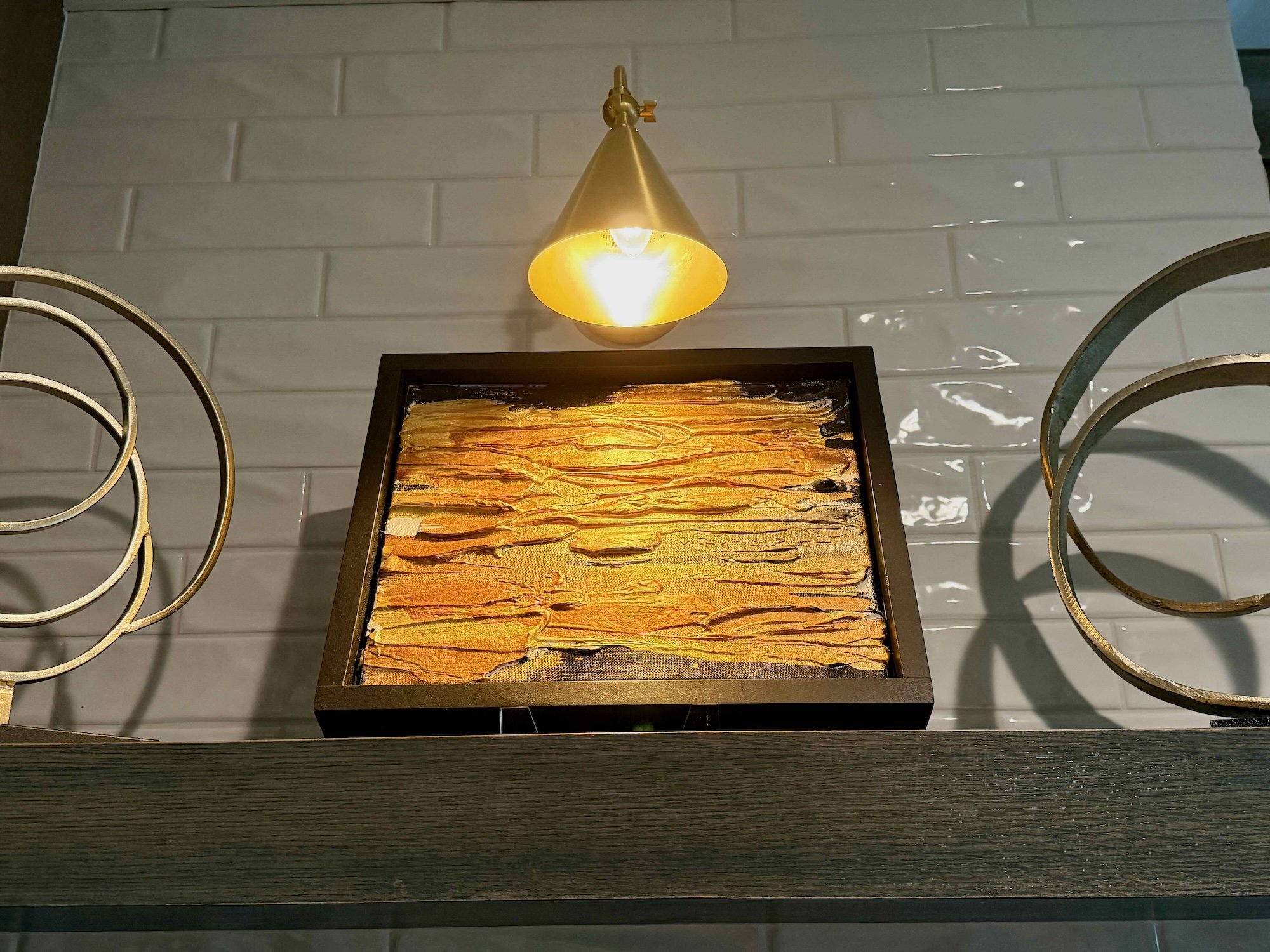 A decorative display with a framed abstract artwork and a wall-mounted gold-colored light fixture above it, on a white tiled wall.