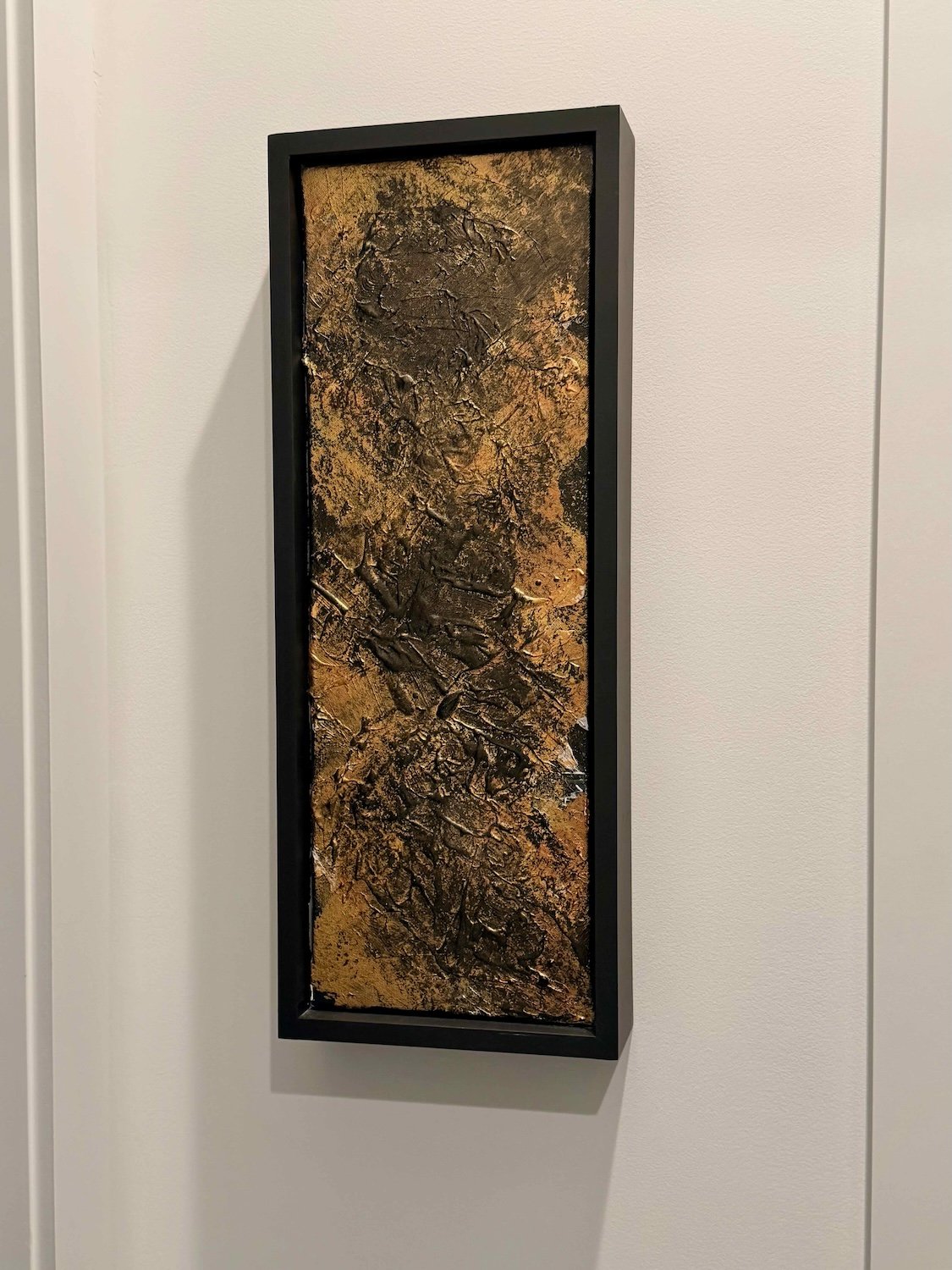 Abstract textured art wall hanging with gold, black, and metallic tones in a black frame on a white wall.