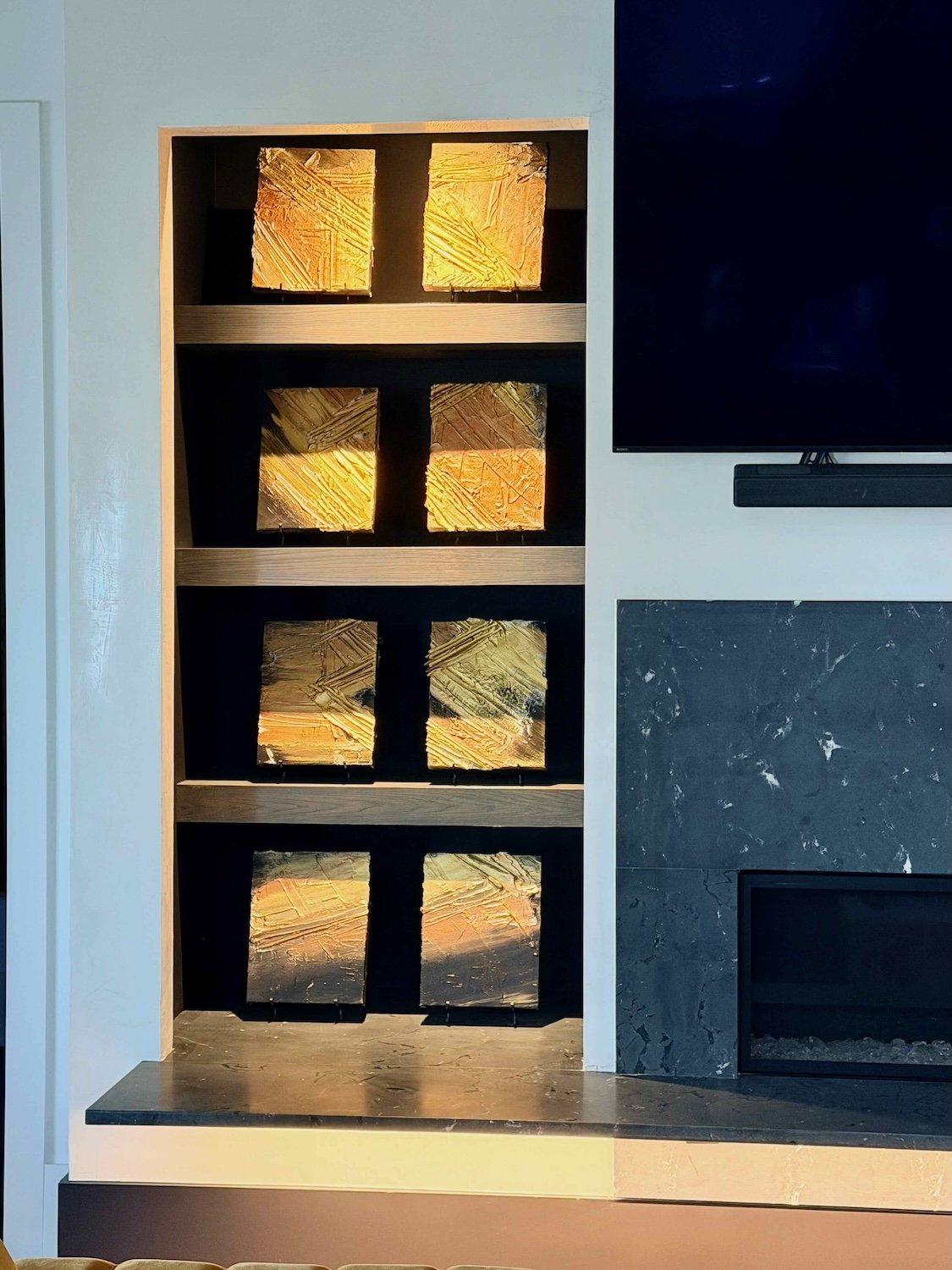 A modern fireplace with a black marble surround and a flat-screen TV mounted above it, with a light-colored wooden shelf and a unique shelving unit to the left featuring illuminated abstract art pieces with textured, colorful surfaces.