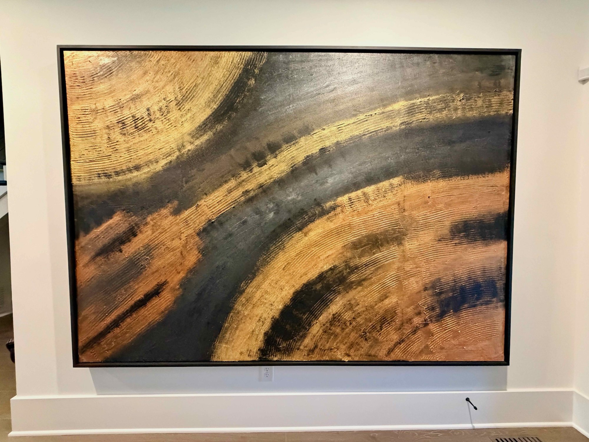 Abstract painting with gold, black, and copper brushstrokes on a white wall.