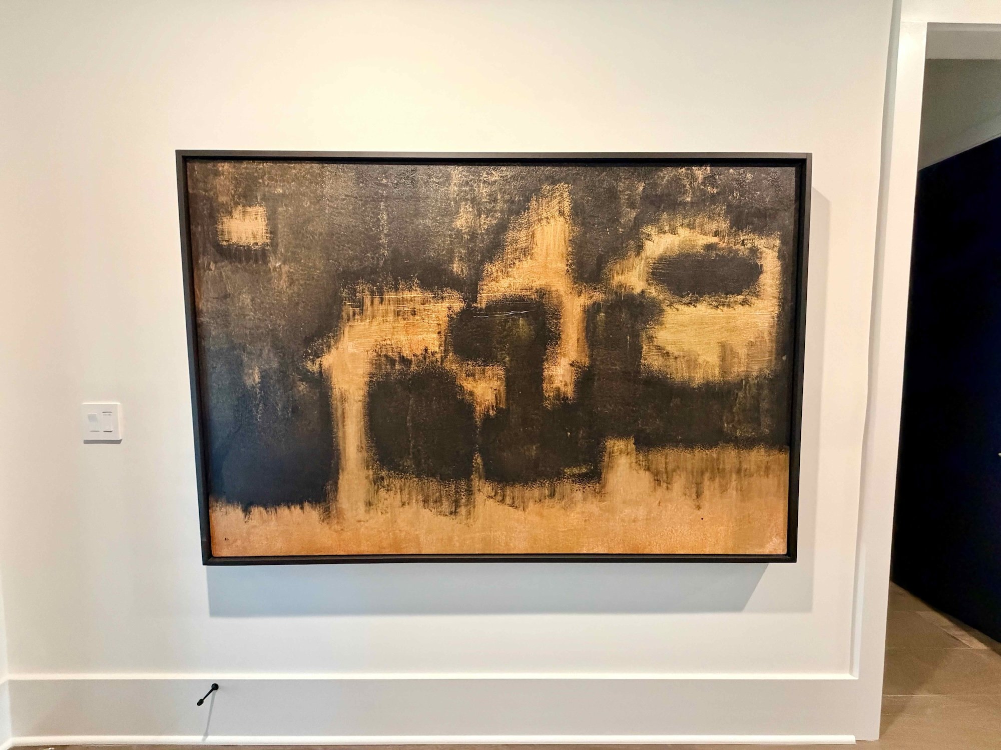 Abstract painting with dark and gold tones mounted on a white wall.