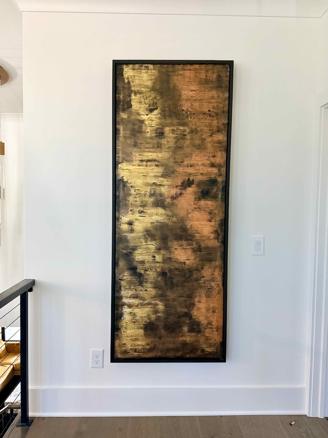 Vertical abstract artwork with blended warm earth tones and dark patches on a white wall.