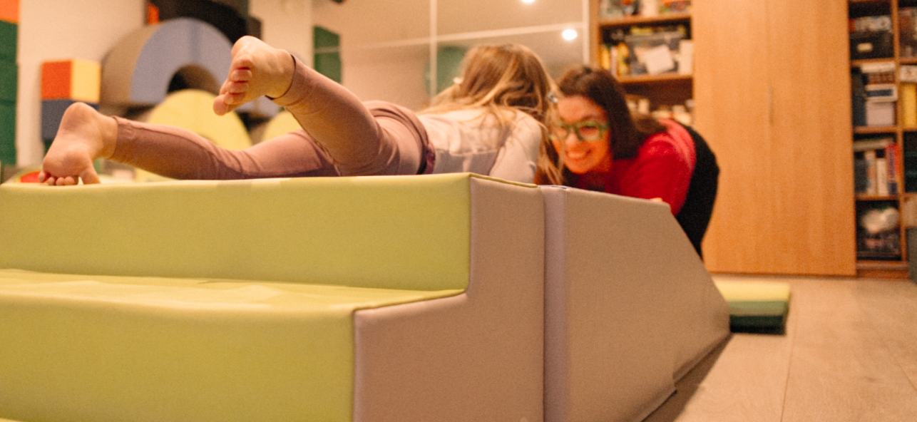 Une jeune fille joue sur un tapis de jeux en intérieur avec une femme, dans une pièce chaleureuse avec des étagères remplies de livres.