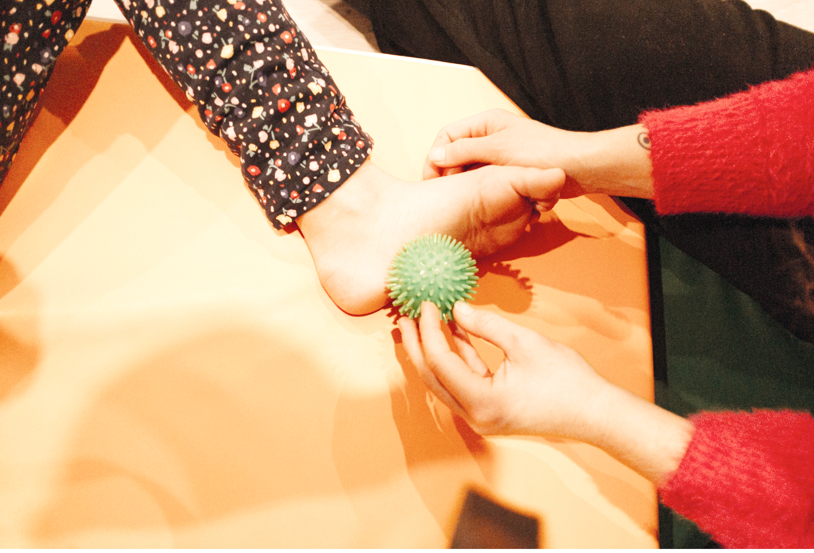 Deux personnes se tiennent la main sur une table orange. Une personne porte un bracelet vert en forme de balle à épines. L'autre personne porte un pull rouge avec des manches en tricot. La scène semble être dans un environnement intérieur.