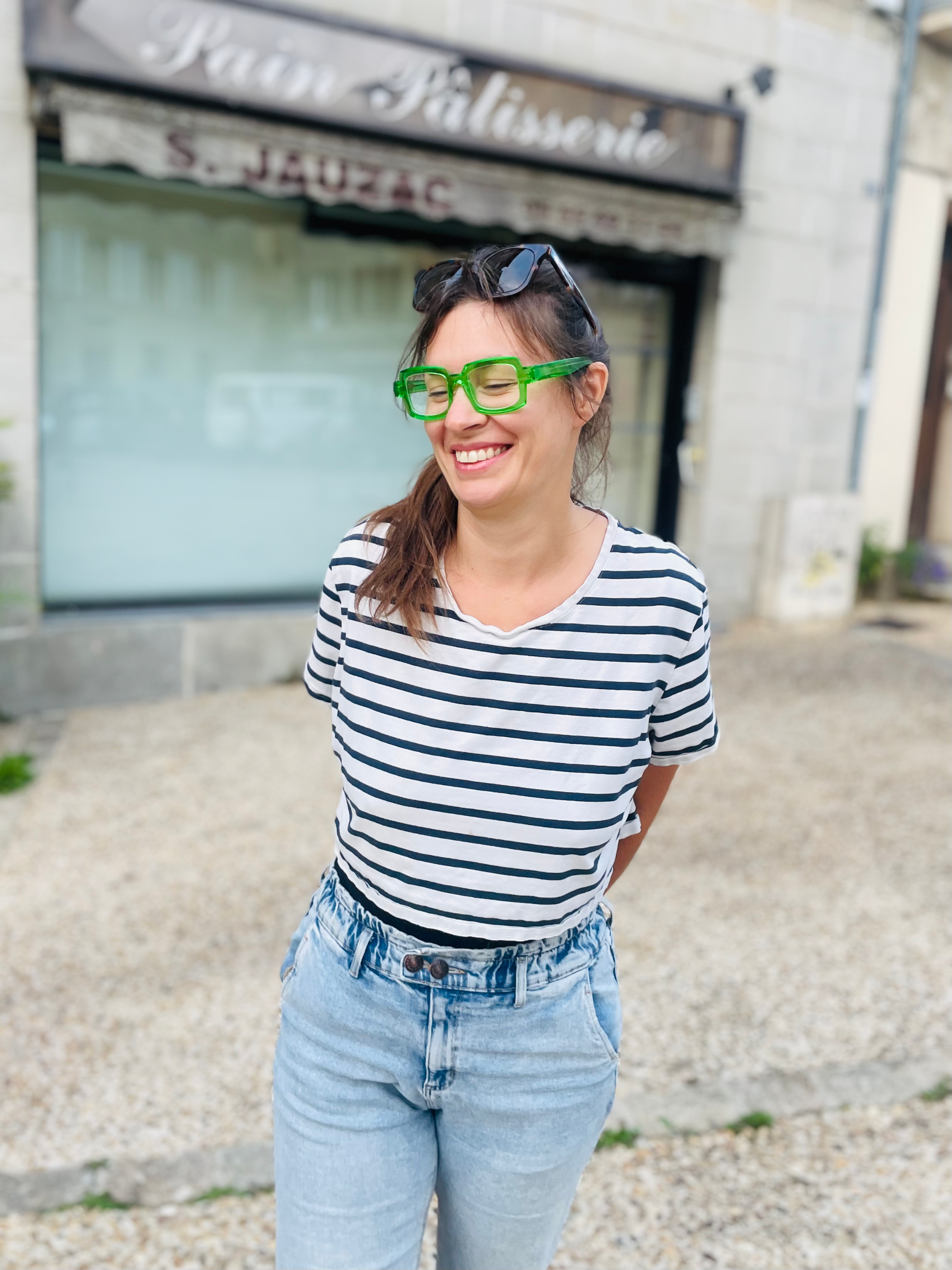 Une femme souriante portant une chemise à rayures et des lunettes vertes, debout devant un bâtiment en pierre.
