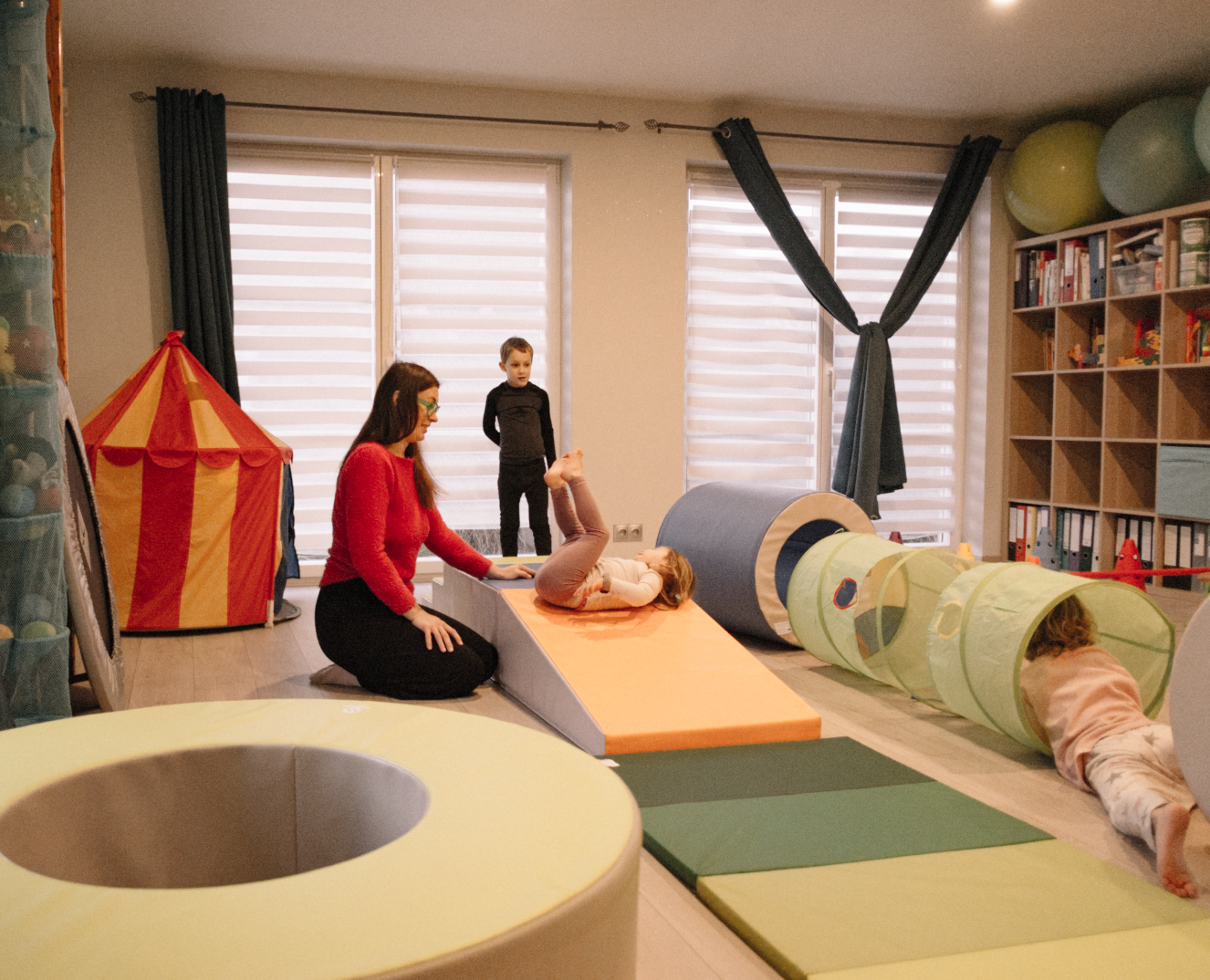 Une salle de jeux pour enfants avec deux adultes et deux enfants. Un enfant court dans une tente en forme de tunnel, un autre enfant et une femme sont sur un tapis dans une zone de jeu. Il y a des coussins, une bibliothèque avec des jouets, des ballons et une tente colorée en arrière-plan.