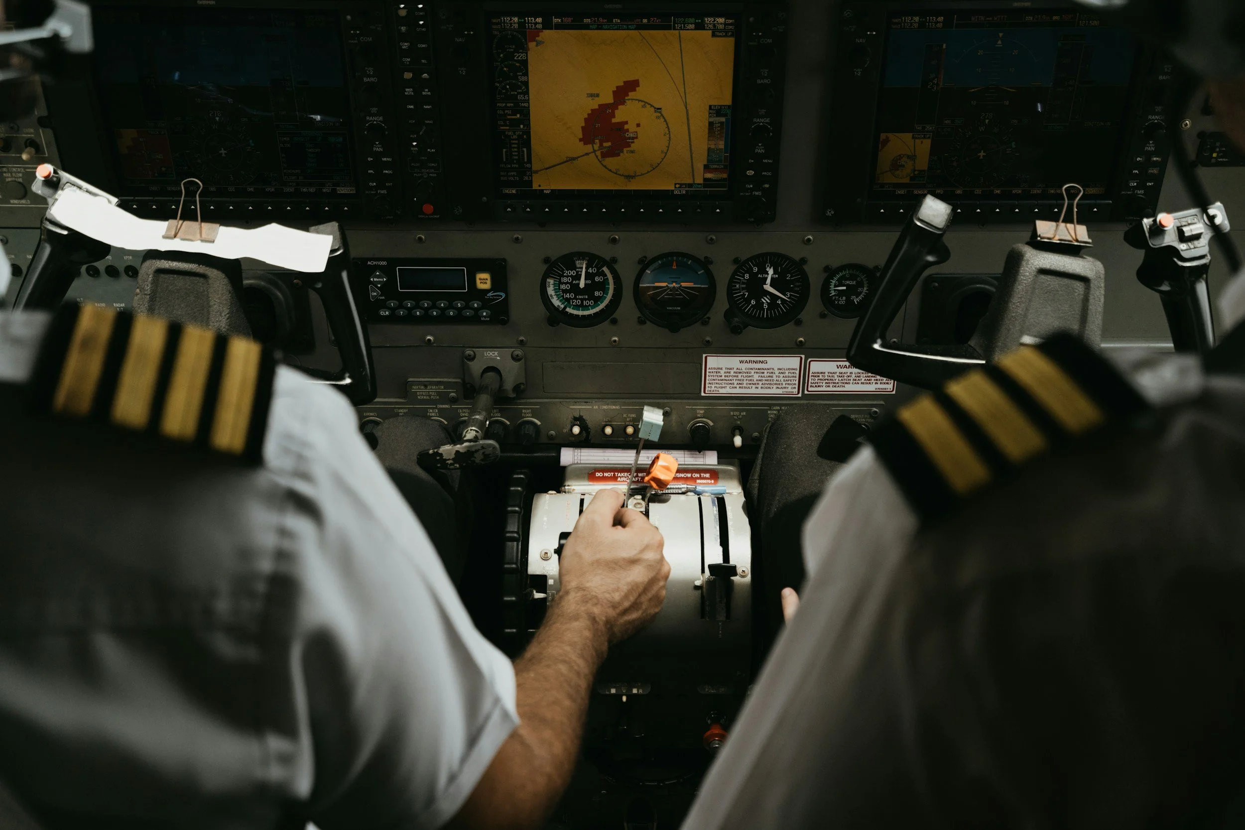 The cockpit of an aircraft, showing two pilots from behind, with various controls and digital screens on the dashboard.