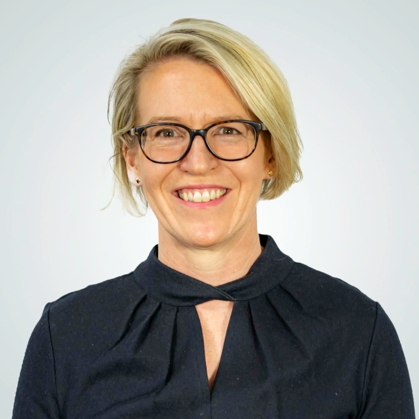 A woman with short blond hair, glasses, and earrings smiling at the camera, wearing a dark top with a pleated neckline.