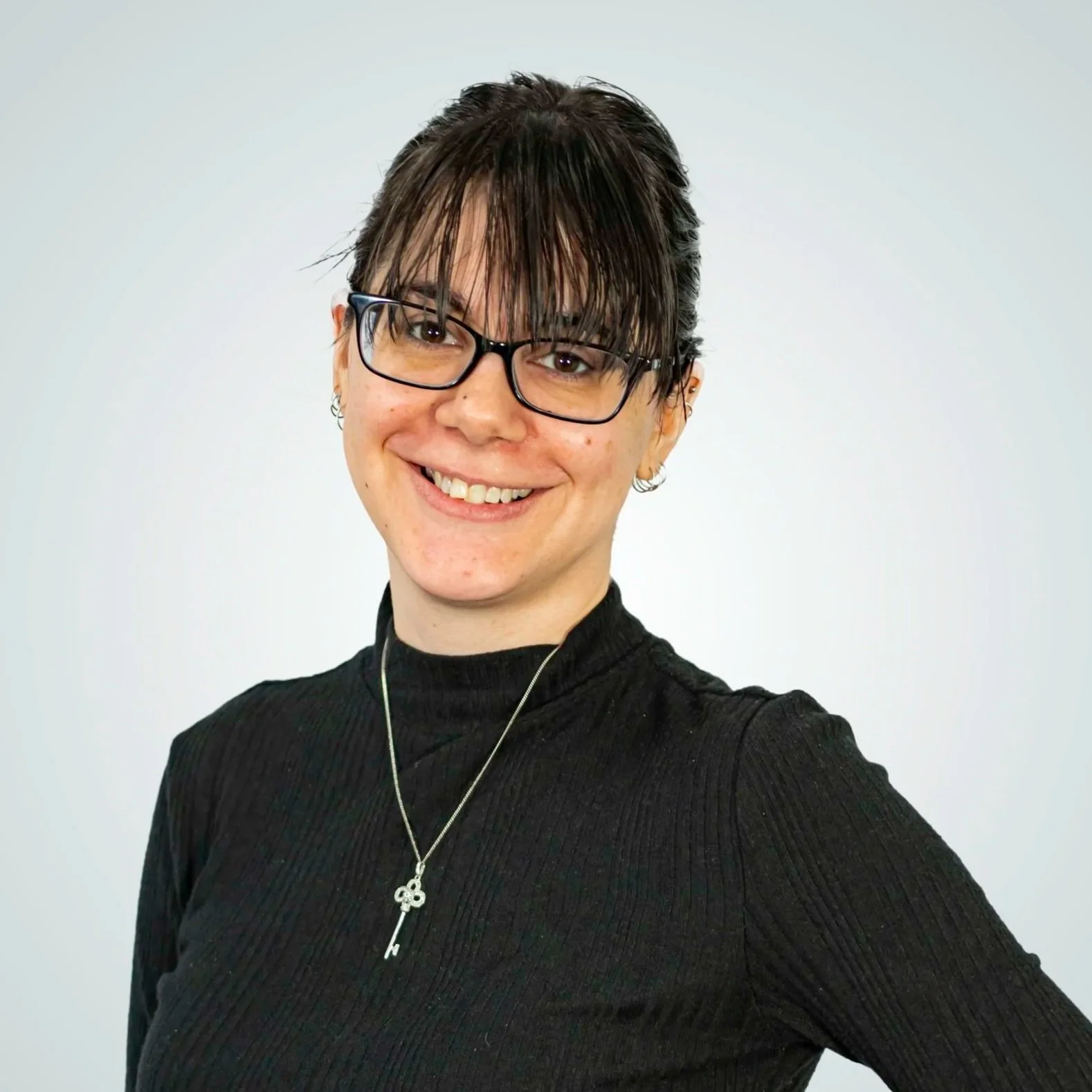 A woman with short dark hair and glasses smiling, wearing a black high-neck shirt and a necklace with a key pendant, standing against a plain background.