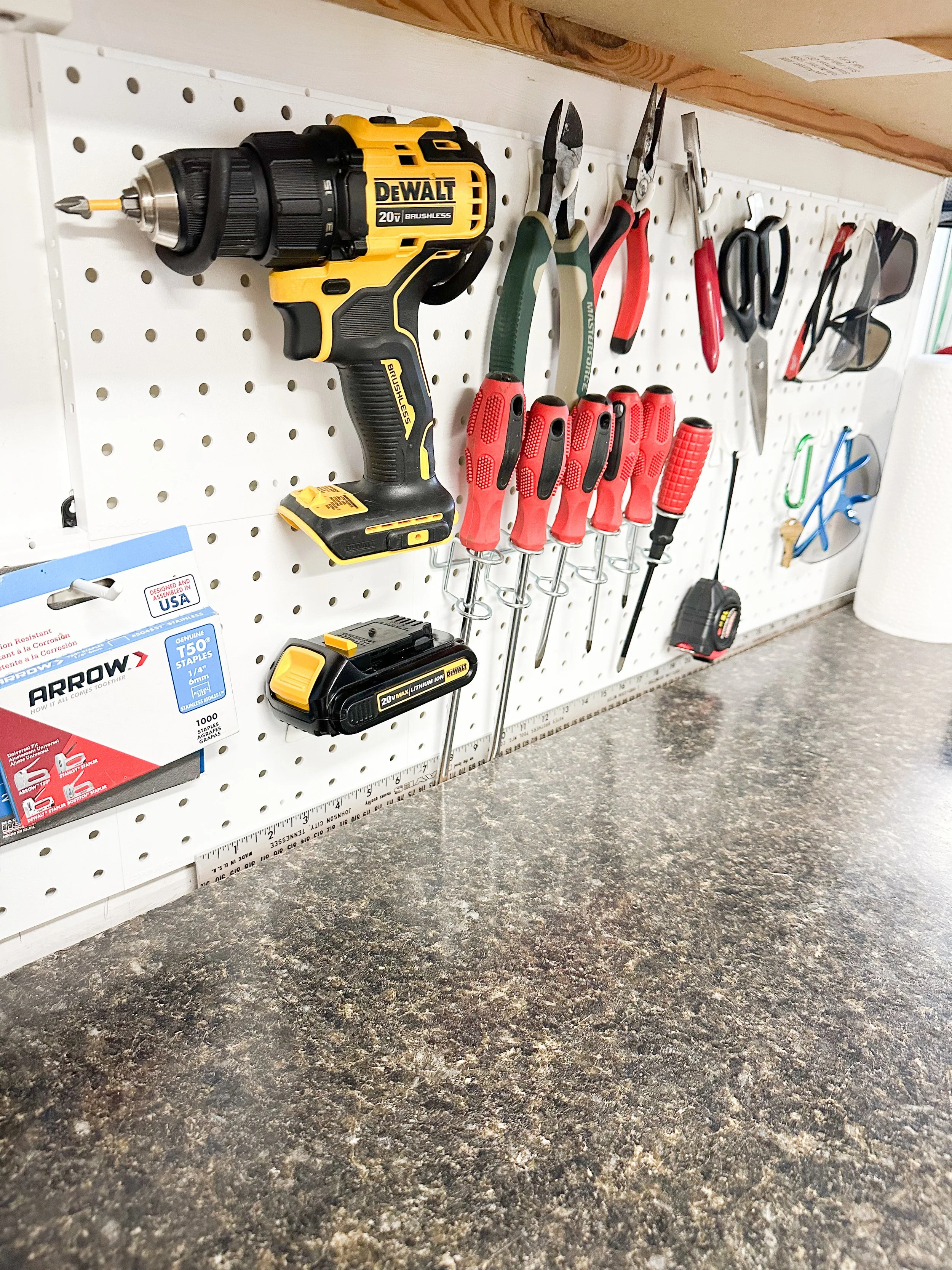 A well-organized pegboard with a cordless drill, screwdrivers, pliers, scissors, a tape measure, and sunglasses, mounted above a speckled workbench.