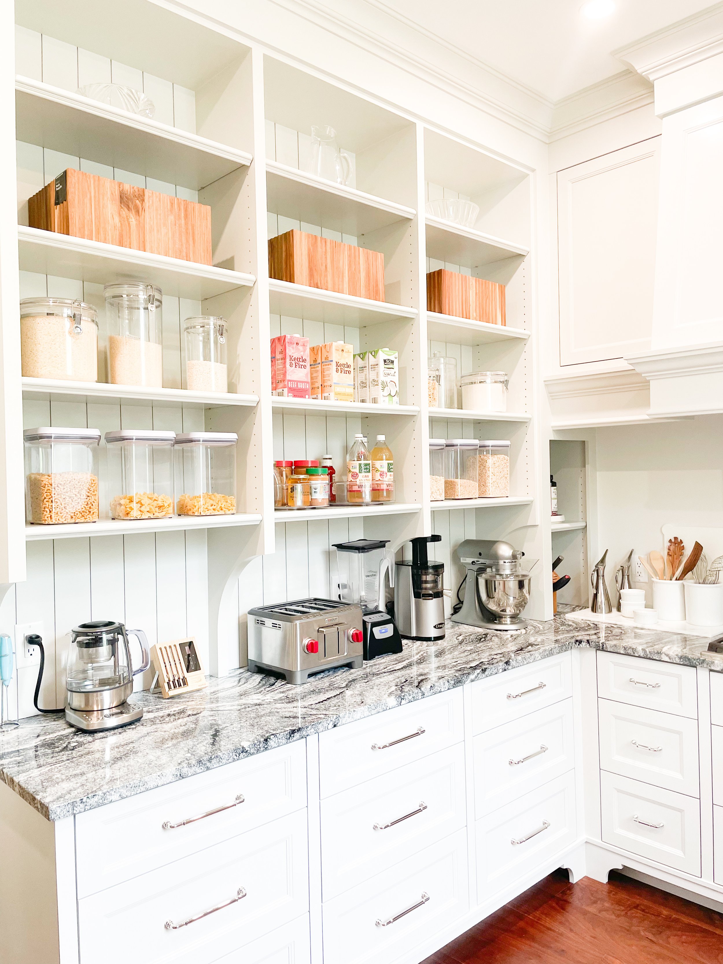White kitchen cabinets with grey marble countertops, open shelves with glass jars, cereal boxes, and decorative wooden boxes, small appliances including a toaster, blender, and stand mixer, in a well-lit space.