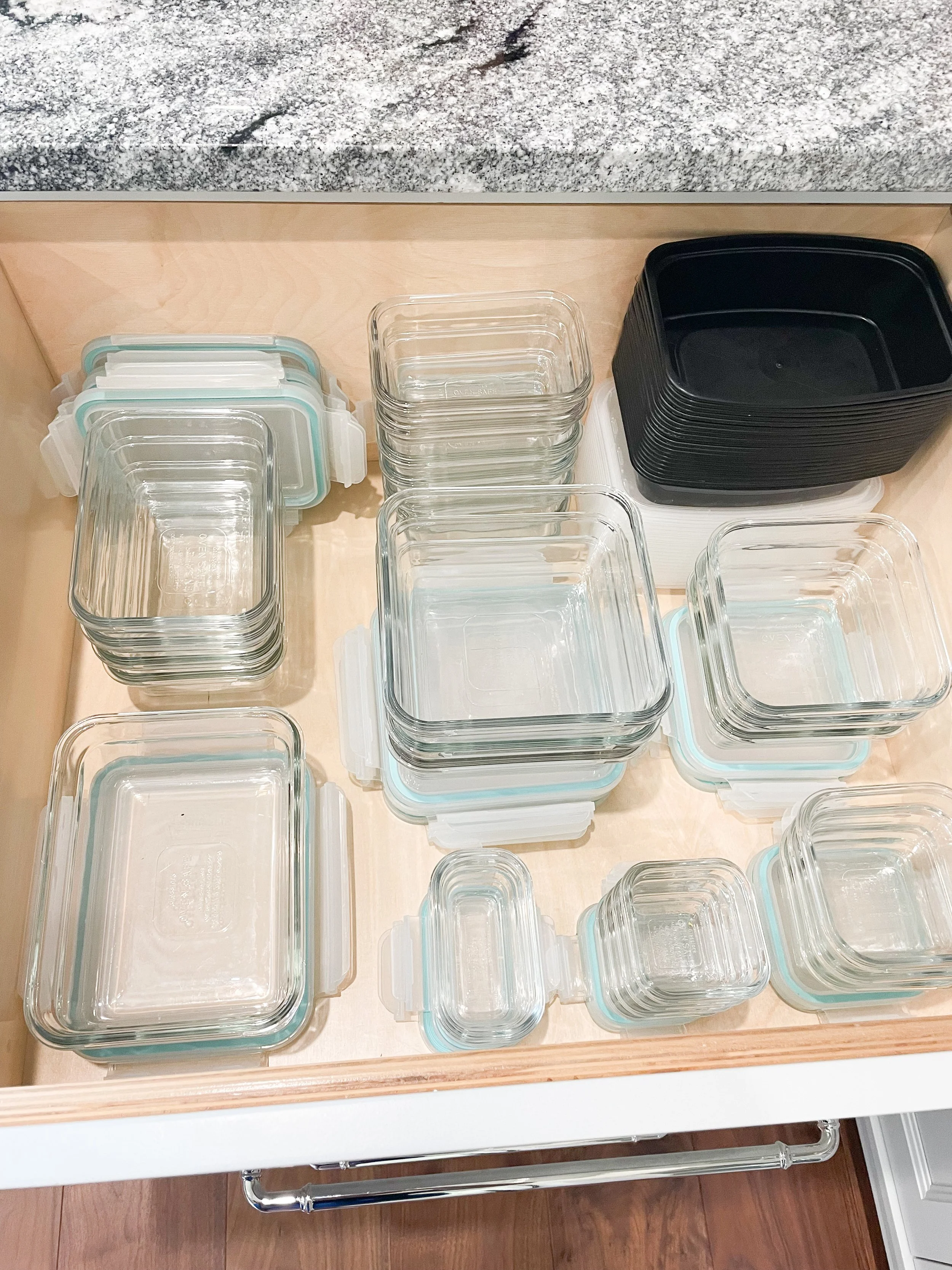 Collection of clear glass and black plastic food storage containers stacked inside a kitchen drawer.