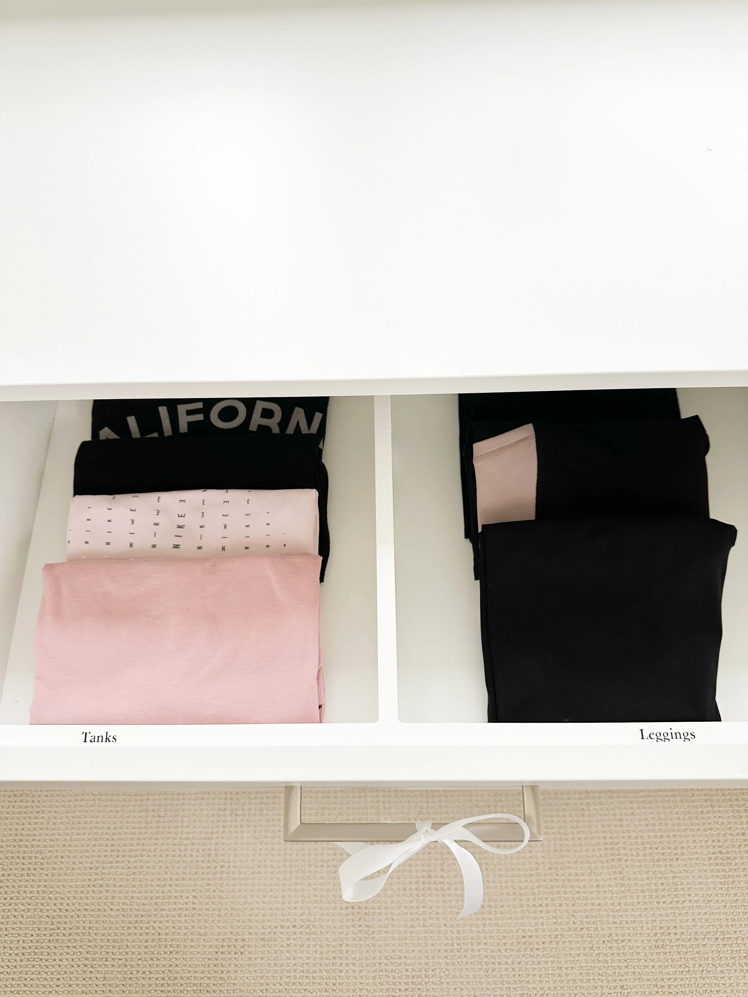 Open white storage drawer containing folded clothing categorized into two sections: left section labeled 'Tanks' with pink, white, and black tanks; right section labeled 'Leggings' with black leggings.