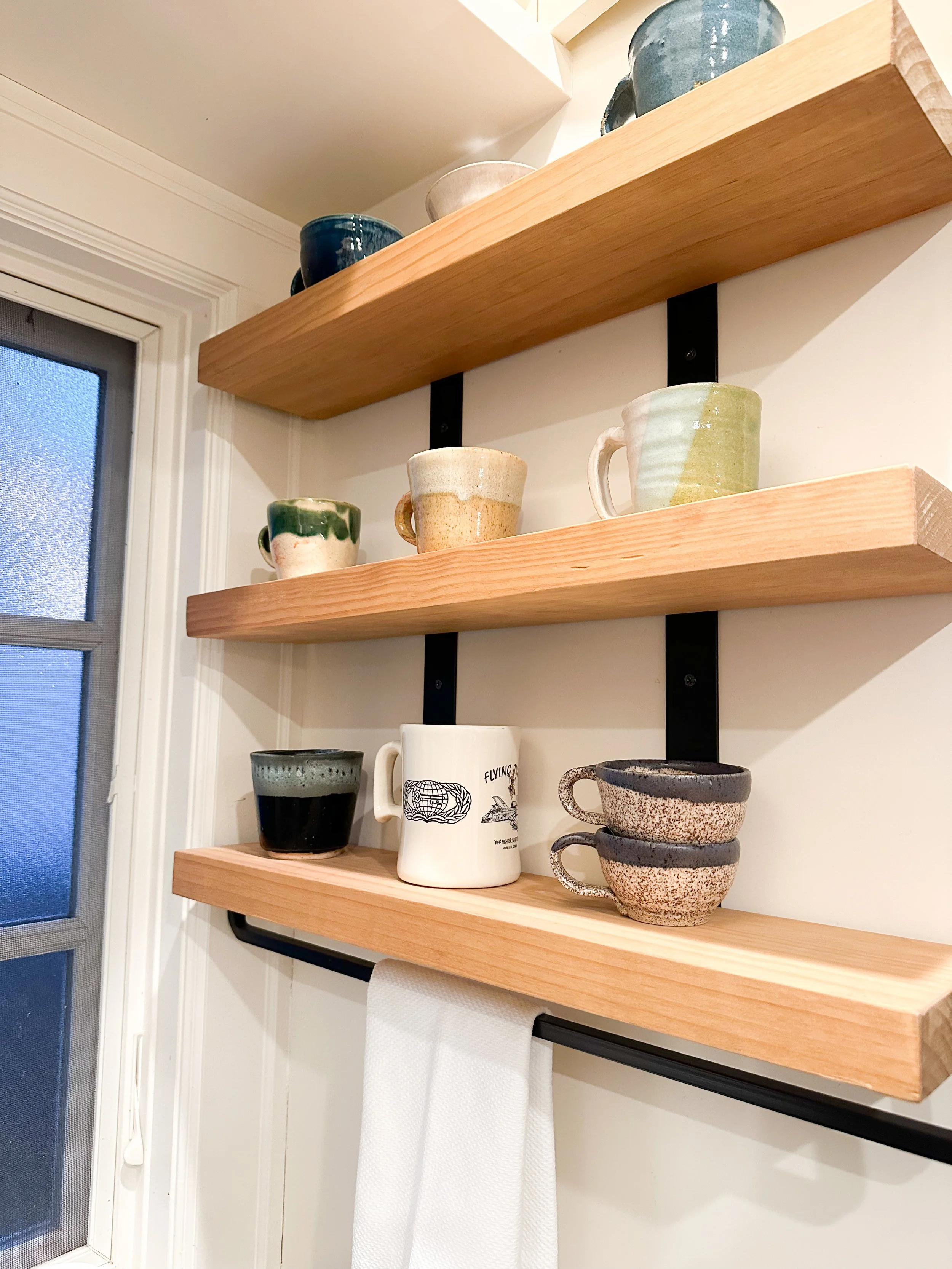 Wooden floating shelves with ceramic mugs and bowls near a window with frosted glass.