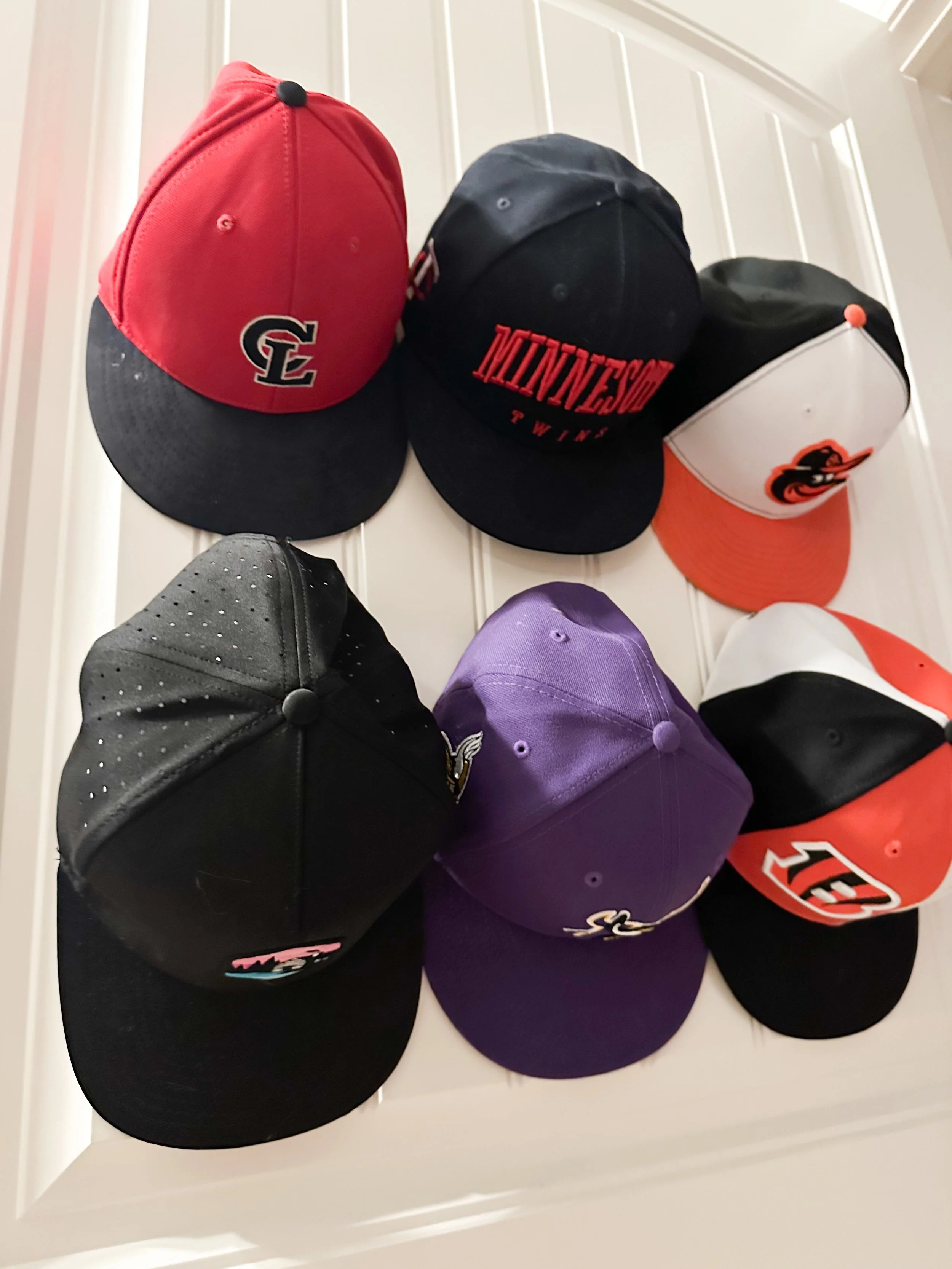 Six baseball caps hanging on a white wall. The caps display various team logos and colors, including red, black, white, purple, and orange.
