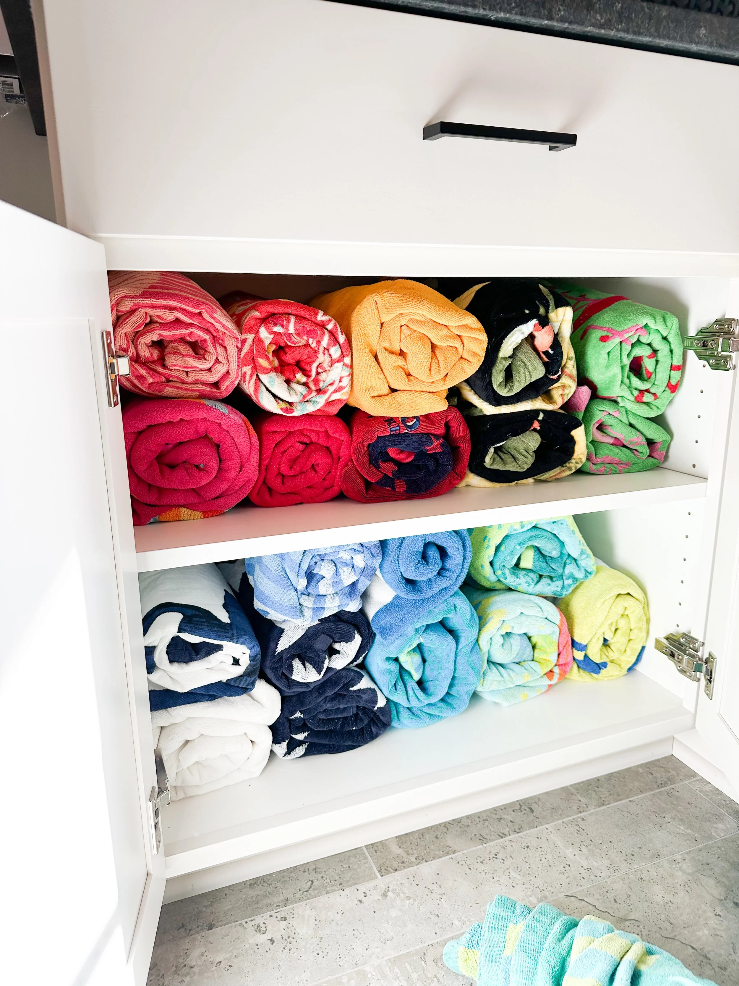 Open white cabinet with three shelves filled with colorful rolled towels. The top shelf has pink, red, yellow, black with floral pattern, and green towels. The middle shelf has white and dark blue towels. The bottom shelf has light blue, dark blue, yellow, and multicolored towels.