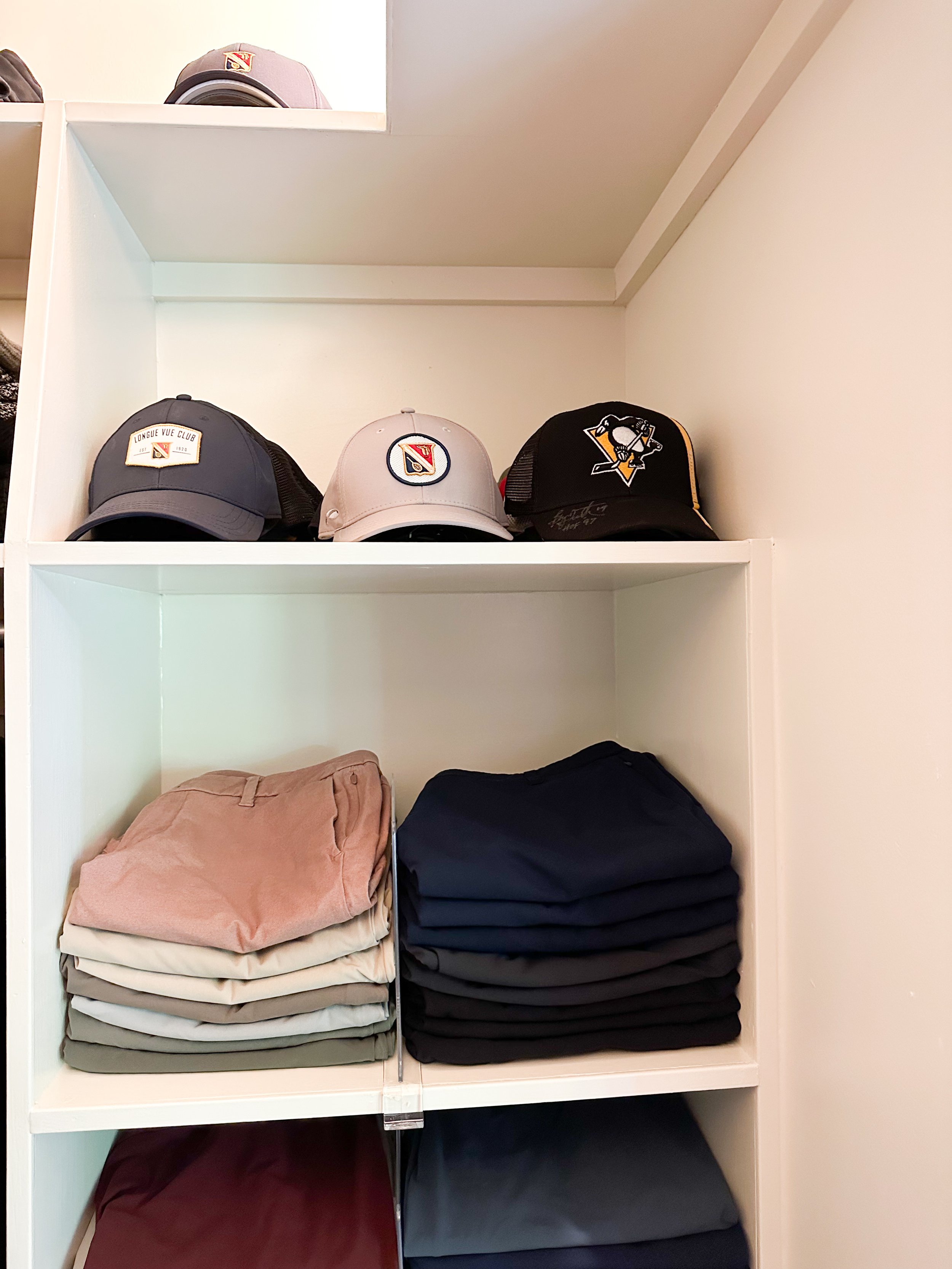 White shelf unit with three hats on the top row featuring sports team logos, and folded clothing in neutral and dark colors on the lower shelves.
