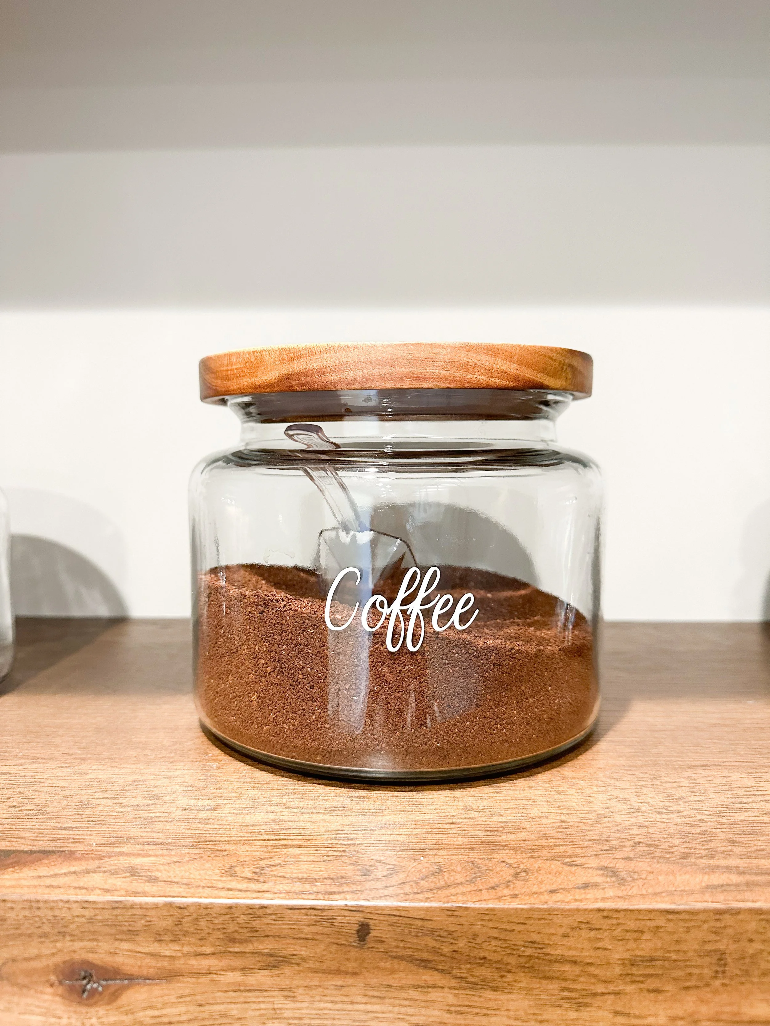 Clear glass jar with a wooden lid labeled 'Coffee' containing ground coffee, placed on a wooden shelf.