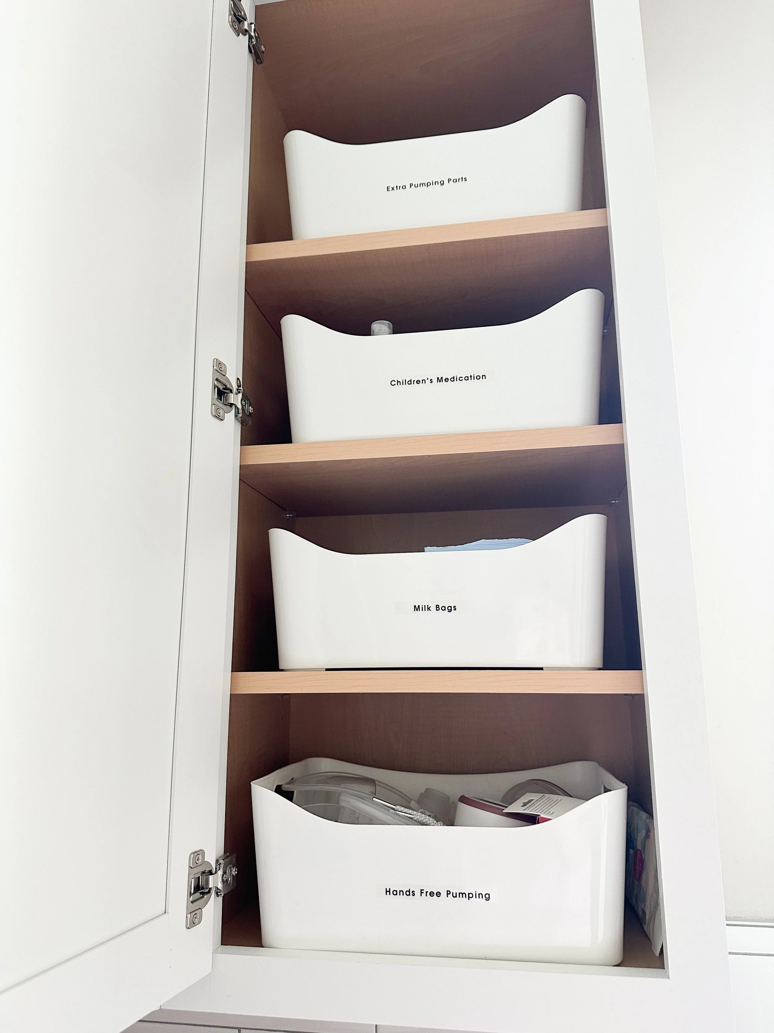 Organized cabinet with four white storage bins labeled 'Extra Pumping Parts', 'Children's Medication', 'Milk Bags', and 'Hands Free Pumping'