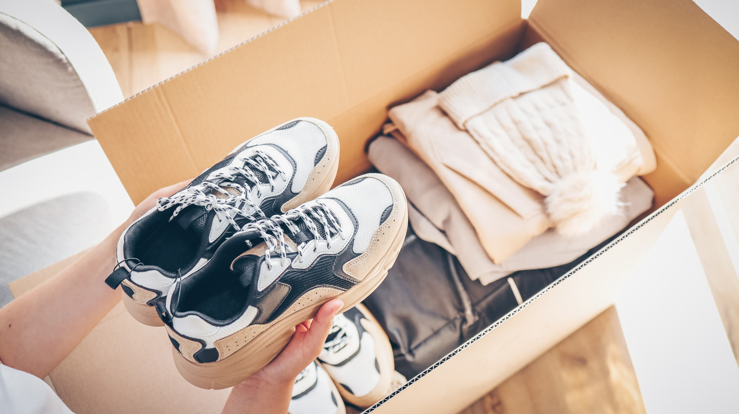 Person holding white and beige sneakers above an open cardboard box containing folded clothes.