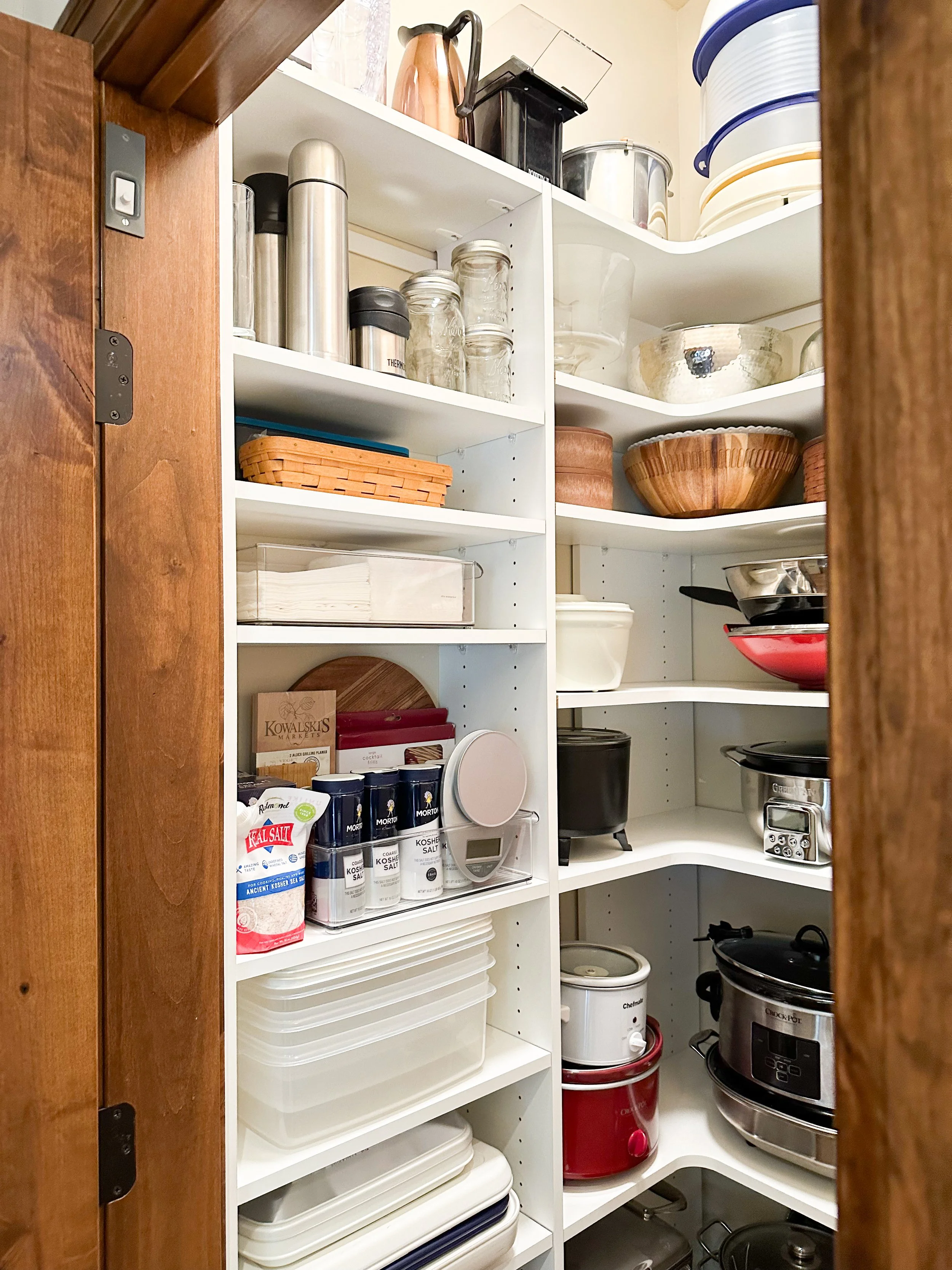 Inside a kitchen pantry with shelves holding glass jars, metal canisters, wooden bowls, plastic containers, a rice cooker, and a slow cooker.