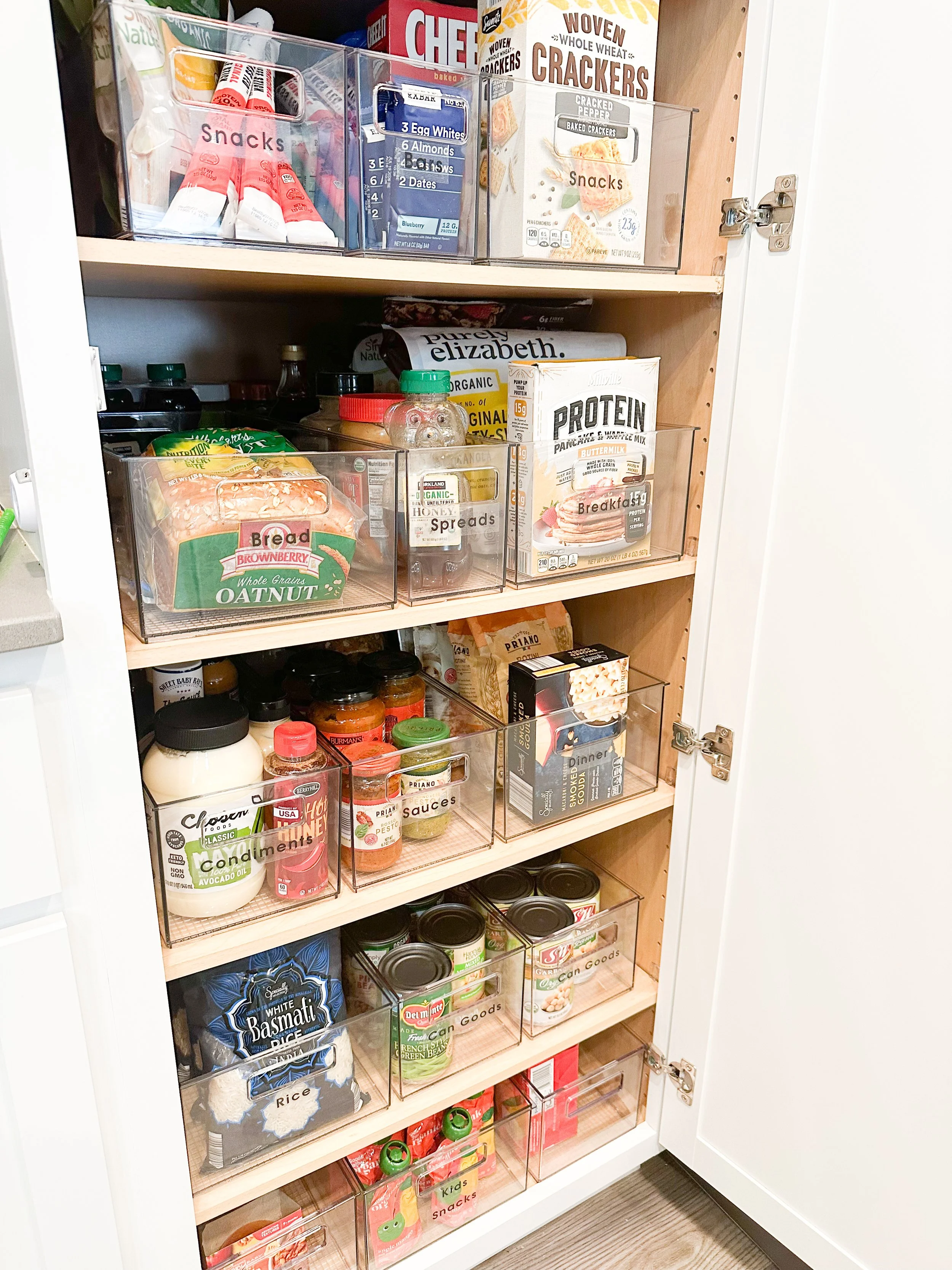 Pantry cabinet with various food storage bins containing snacks, cereals, canned goods, sauces, and baking supplies.