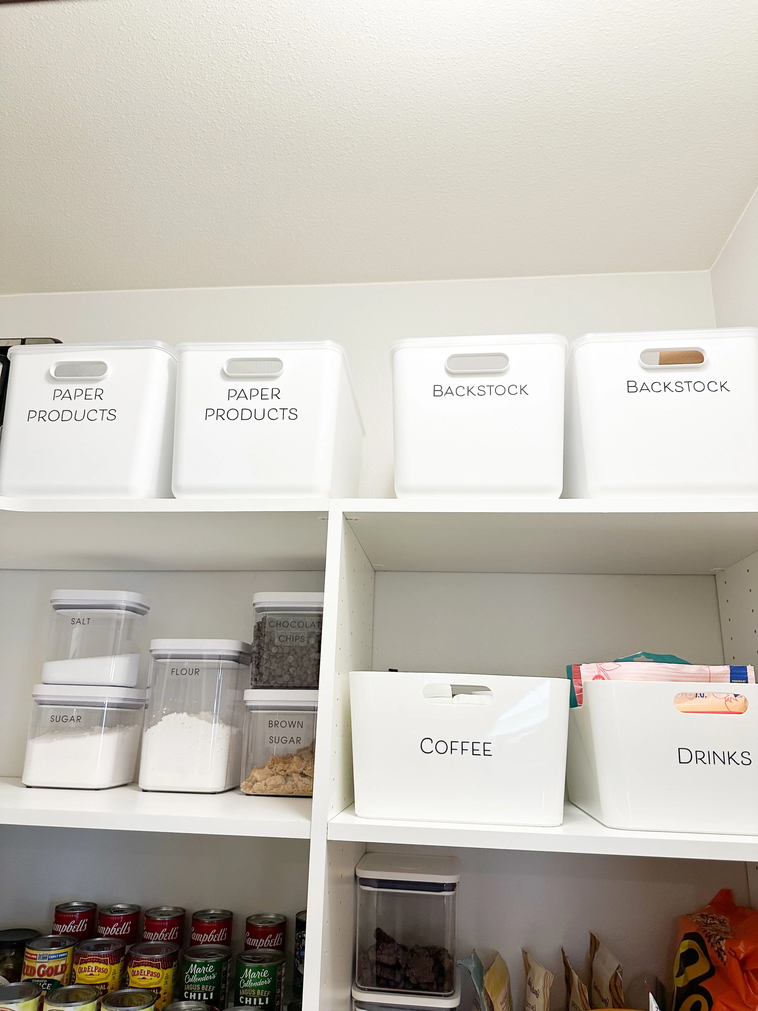 White storage bins labeled 'PAPER PRODUCTS' and 'BACKSTOCK' on a white kitchen shelf. Smaller containers labeled 'SALT', 'FLOUR', 'SUGAR', 'BROWN SUGAR', and 'CHOCOLATE CHIPS' below. A white container labeled 'COFFEE' and another labeled 'DRINKS' with snacks and food items below.