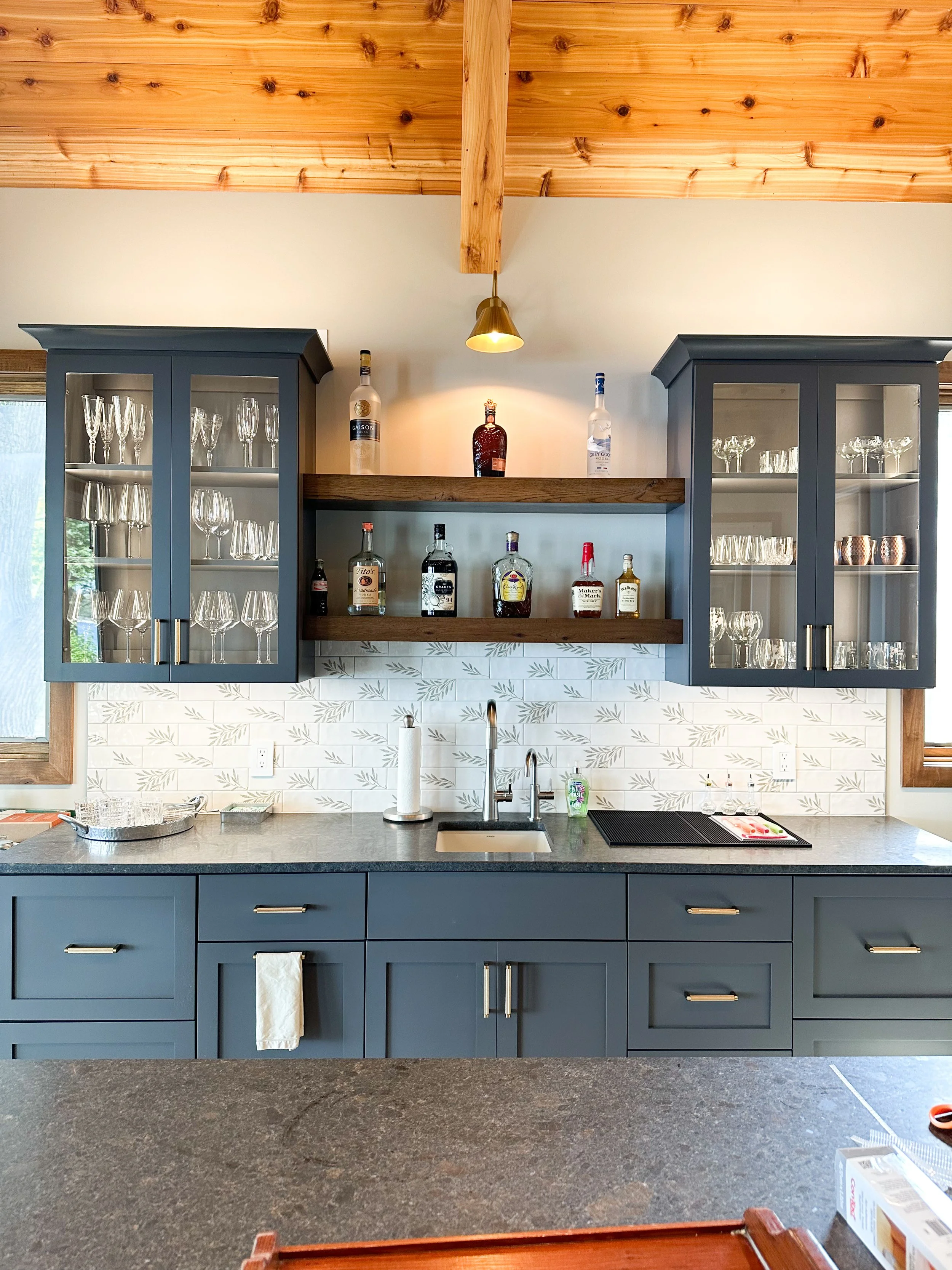Kitchen with blue cabinets, open shelves with glasses and bottles, gray countertop, white patterned tile backsplash, wood ceiling, and a window on each side.