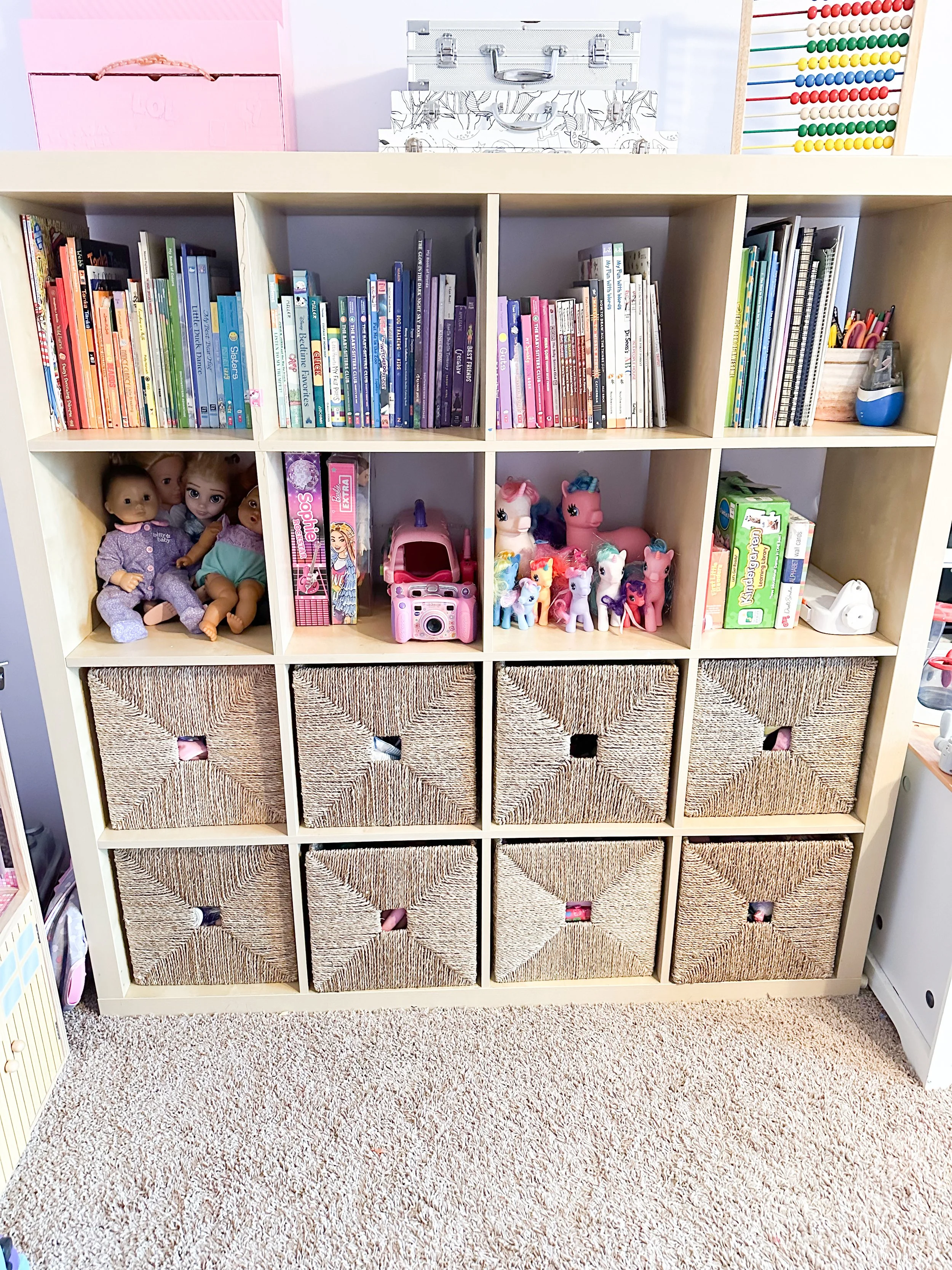 A white cube storage shelf filled with children's toys, books, and dolls, with woven baskets in the bottom row and a carpeted floor at the base of the shelf.